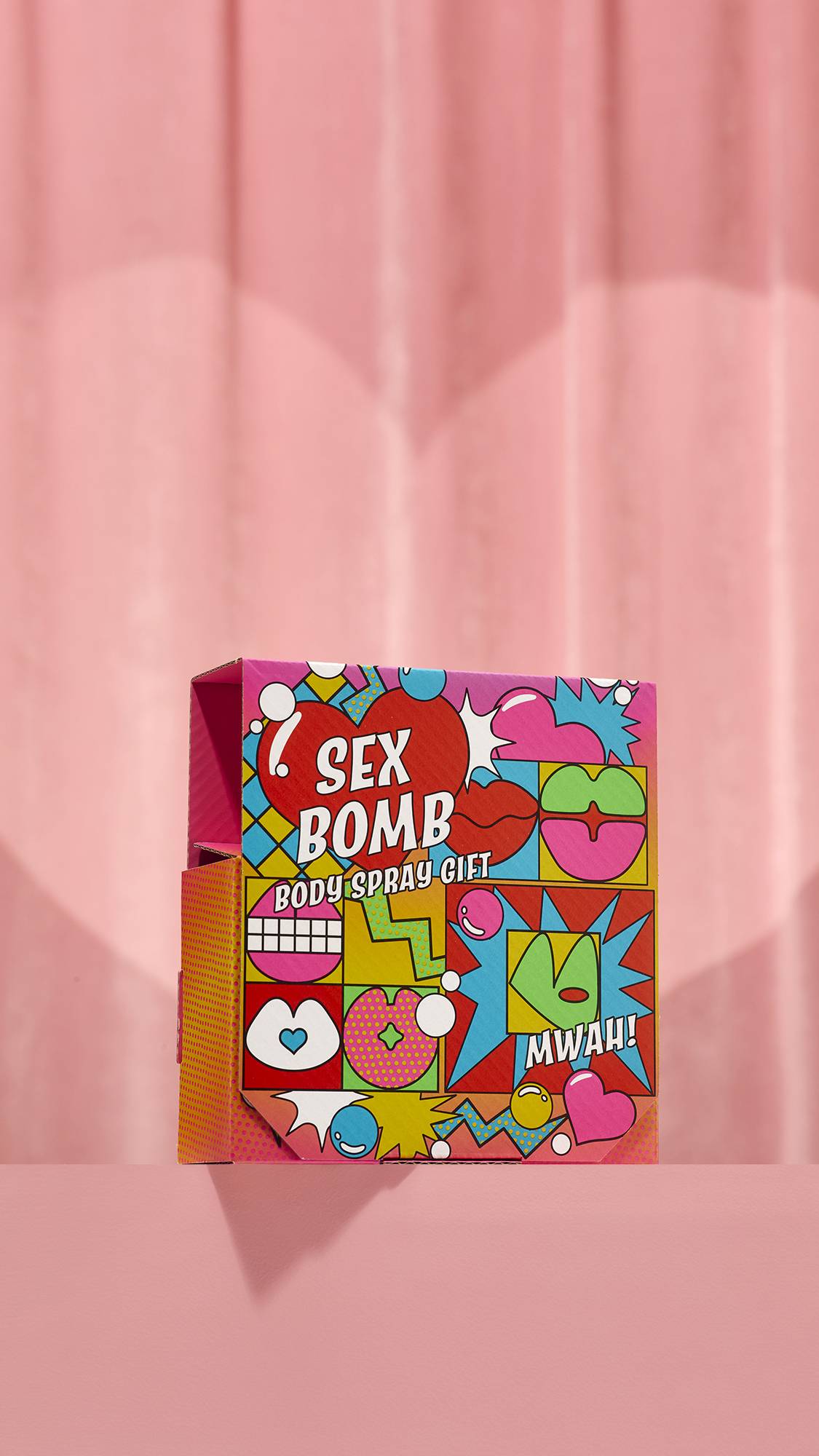 The Sex Bomb Body Spray Gift features a vibrant pop-art box decorated with illustrations of red hearts, colorful lips, and speech bubbles reading Mwah. It sits on a pink pedestal against a soft pink curtain background.
