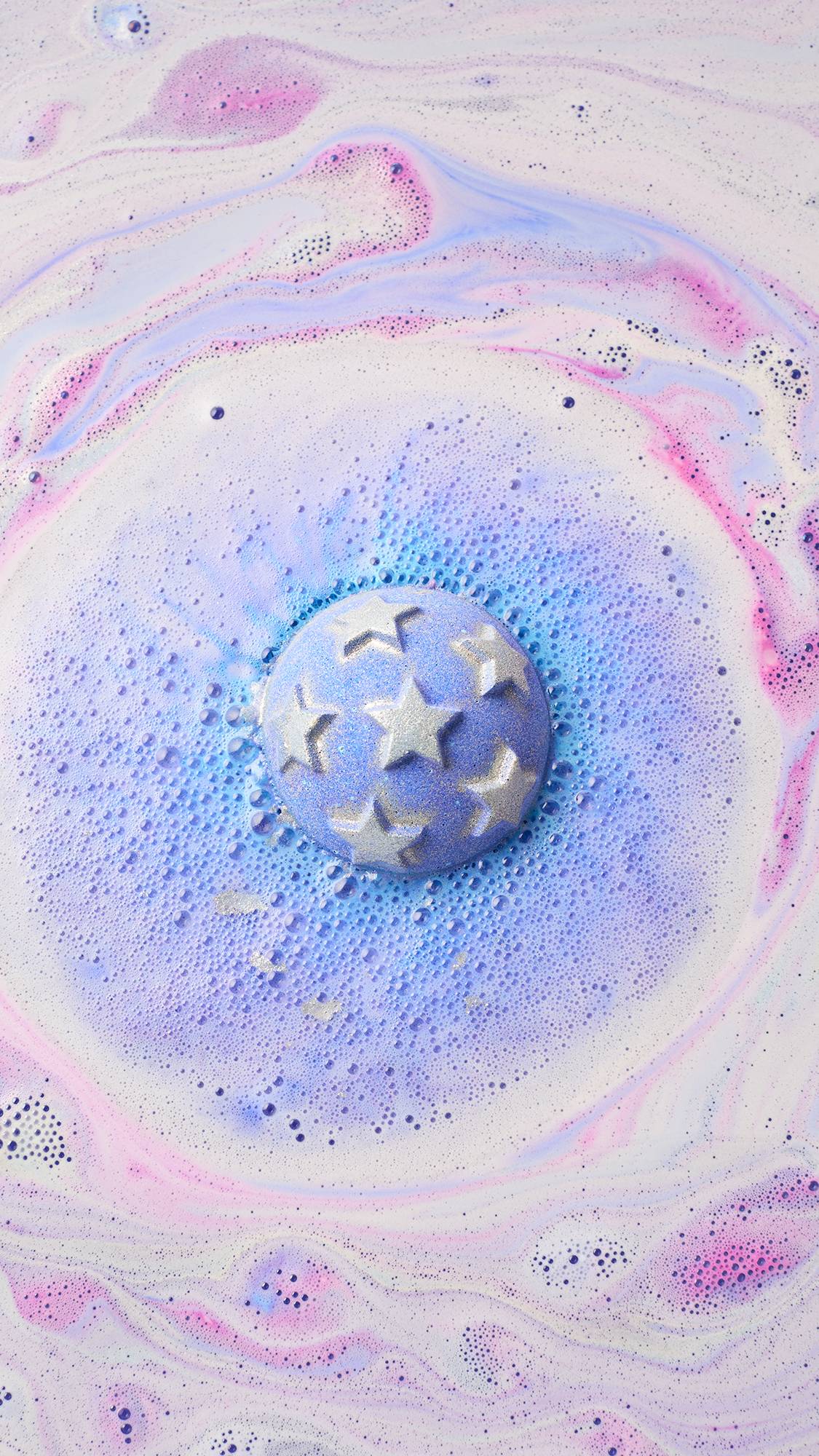 A spherical purple bath bomb embossed with silver-white stars dissolves in bathwater, expanding into a ring of frothy blue fizz surrounded by swirls of pastel pink and white foam.