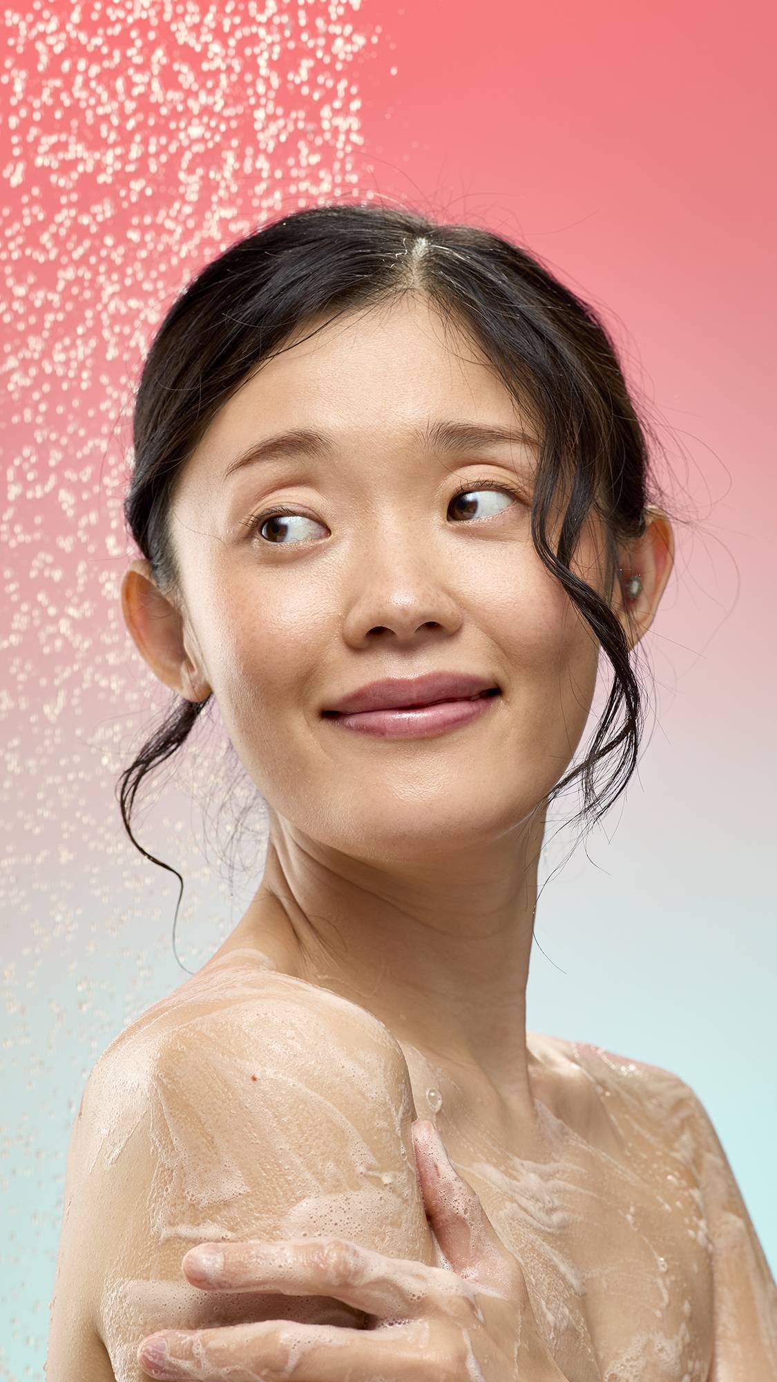 A person smiles while applying a foamy white lather from Smell Of Roses soap to their shoulder. They are standing under a spray of falling water droplets against a soft pink-to-blue gradient background.