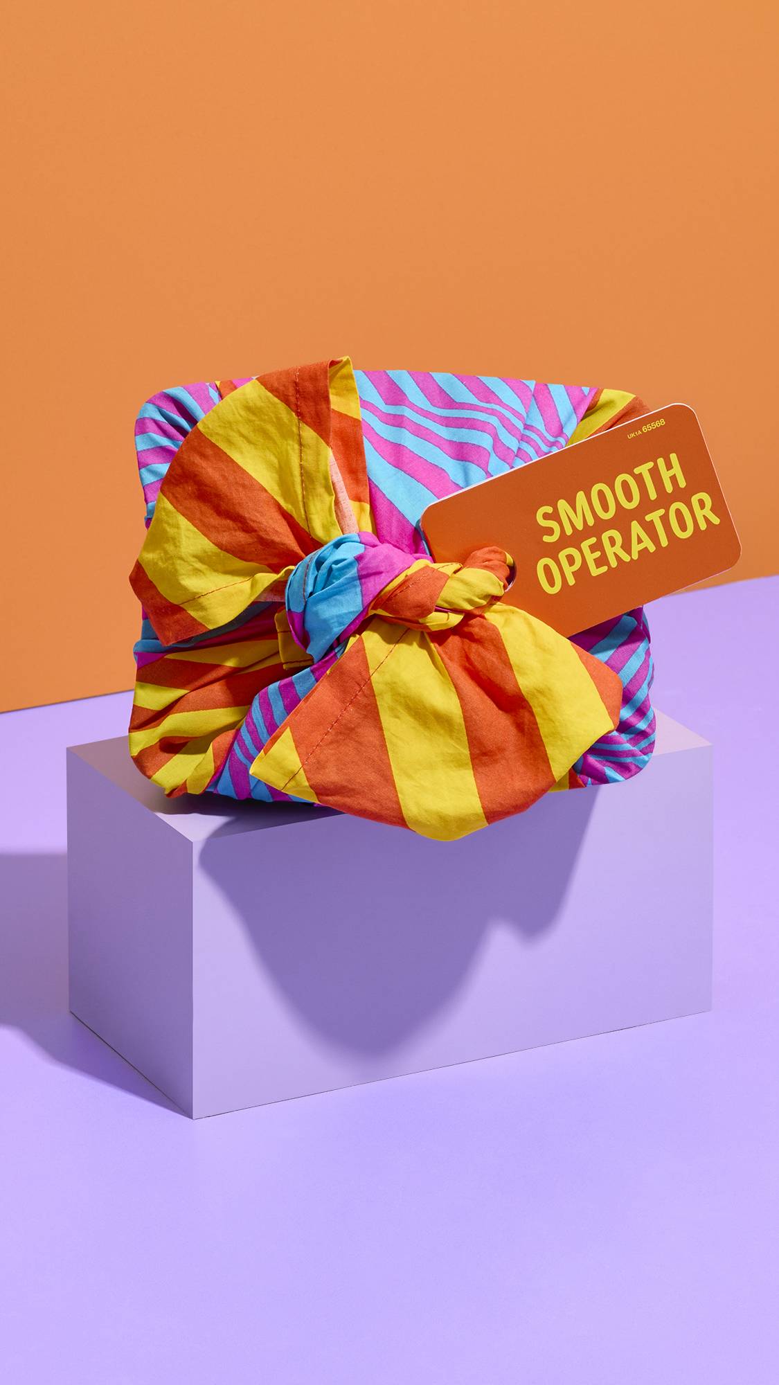 A brightly patterned knot-wrapped gift tagged "SMOOTH OPERATOR" sits on a lilac plinth, featuring a yellow and orange striped knot tied against a split background of orange and purple.
