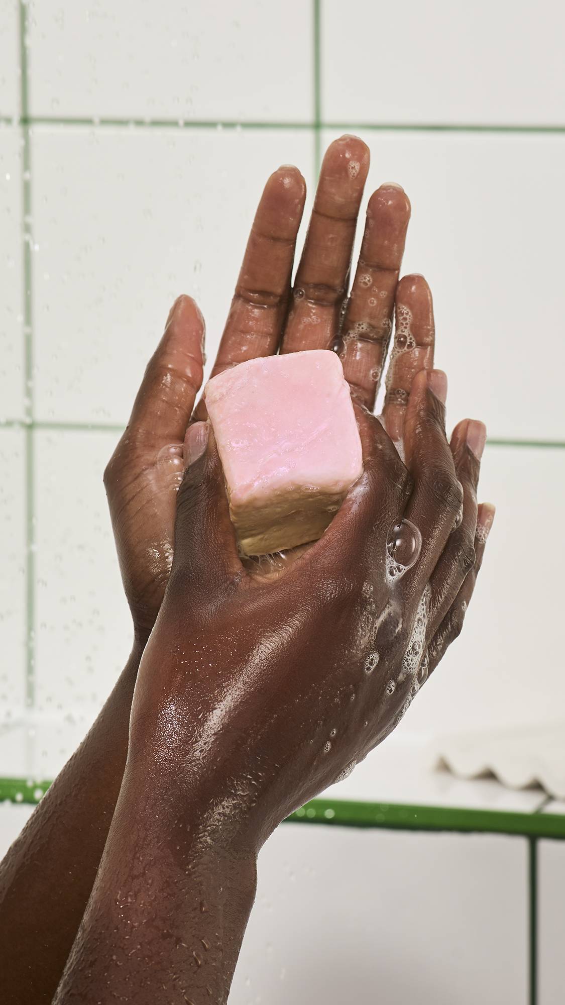 Hands hold a cubed "S'mores" soap with a smooth pink top layer under streaming shower water, building up light white bubbles against a white tiled wall background.