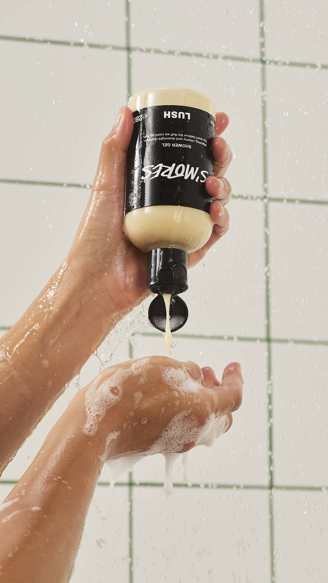 A person squeezes a bottle of Lush "S'mores" shower gel upside down in the shower, pouring a smooth stream of creamy, off-white gel into their soapy hand against a white tiled wall.