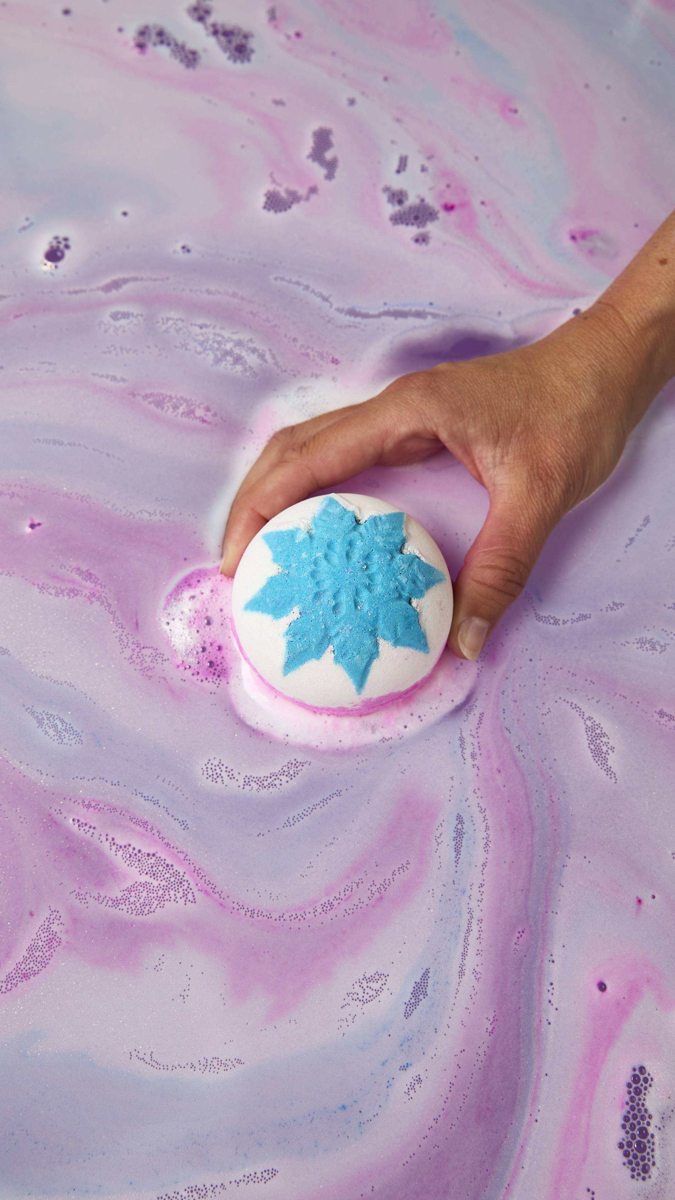 A hand places a white round "Snow Fairy" bath bomb topped with a vibrant blue snowflake onto pastel pink and purple bathwater, releasing a ring of fine pink foam.