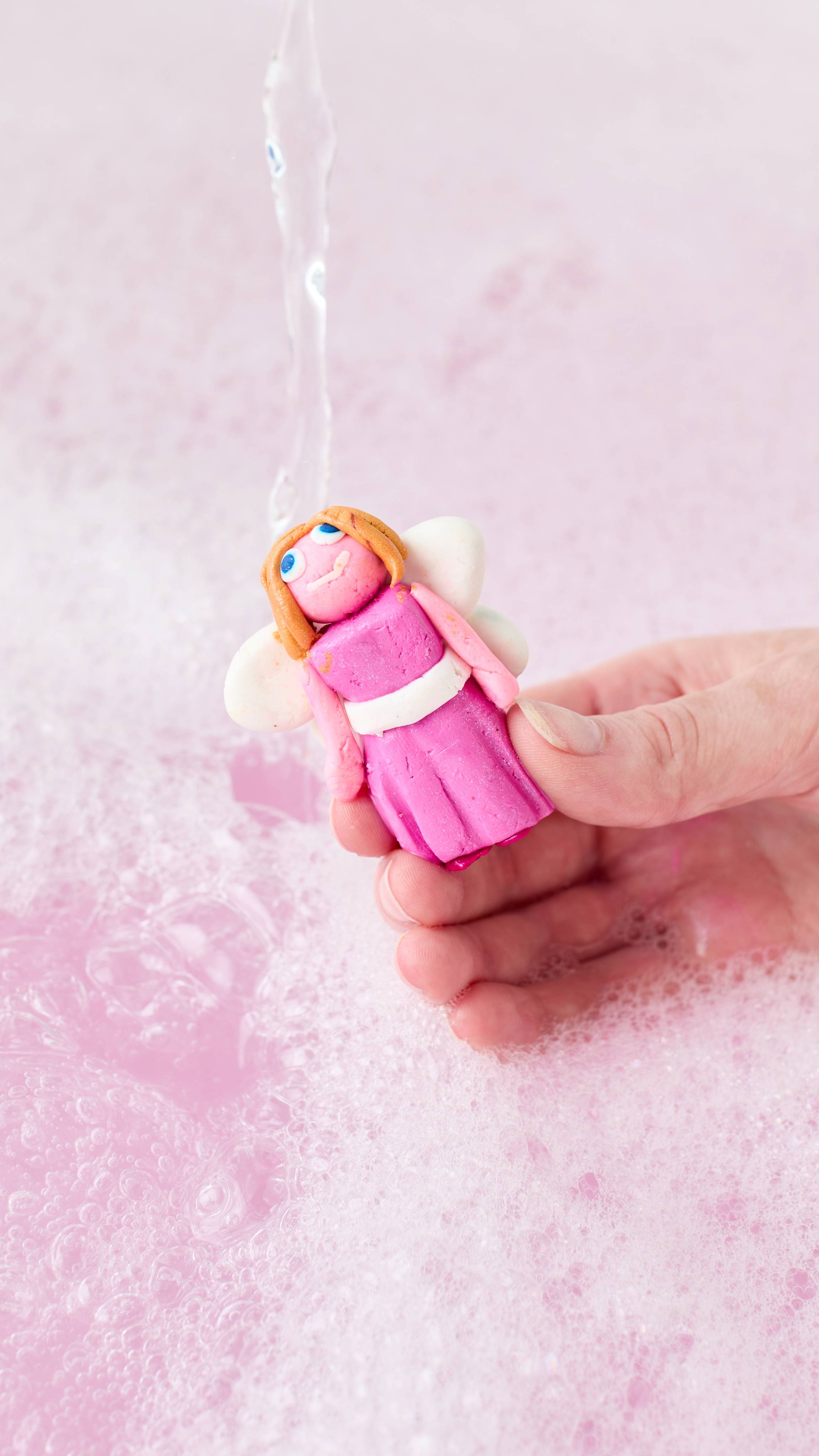 A hand holds a small, colourful fairy-shaped character made from Lush "Snow Fairy" FUN, under a stream of tap water, creating soft pink foam and bubbles in the bathwater below.