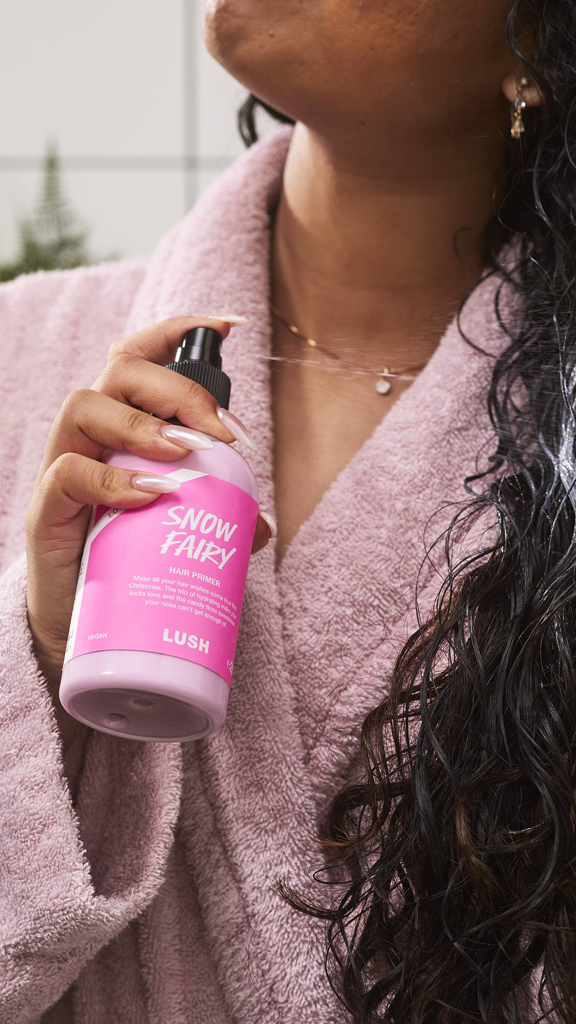 A person in a cosy plush pink dressing gown sprays Lush "Snow Fairy" hair primer onto their long dark curly hair.