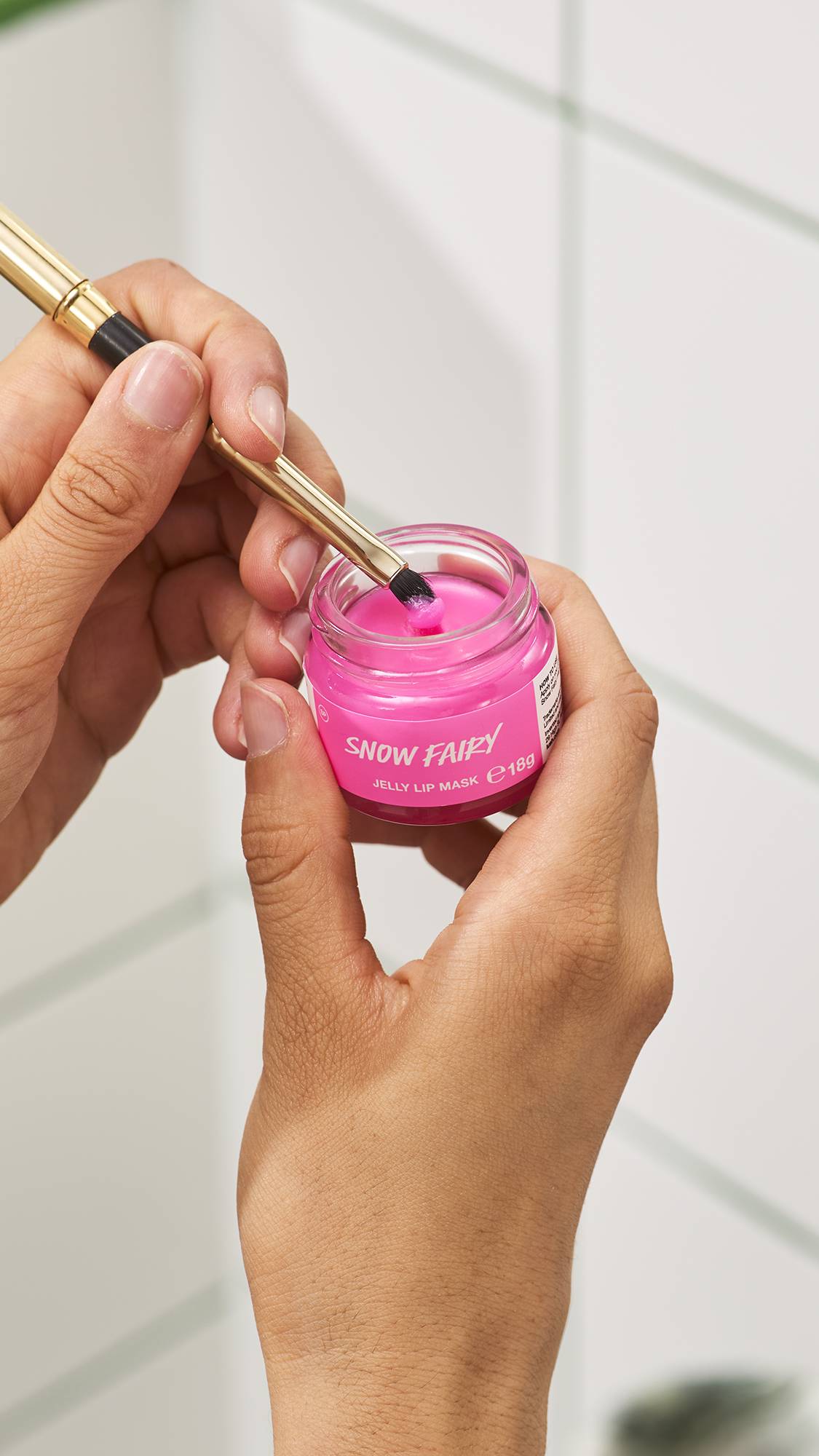 A person holds a small glass jar of vibrant bright pink Lush Snow Fairy jelly lip mask in one hand while using a lip brush to scoop product from it, set against a white tiled bathroom wall background.