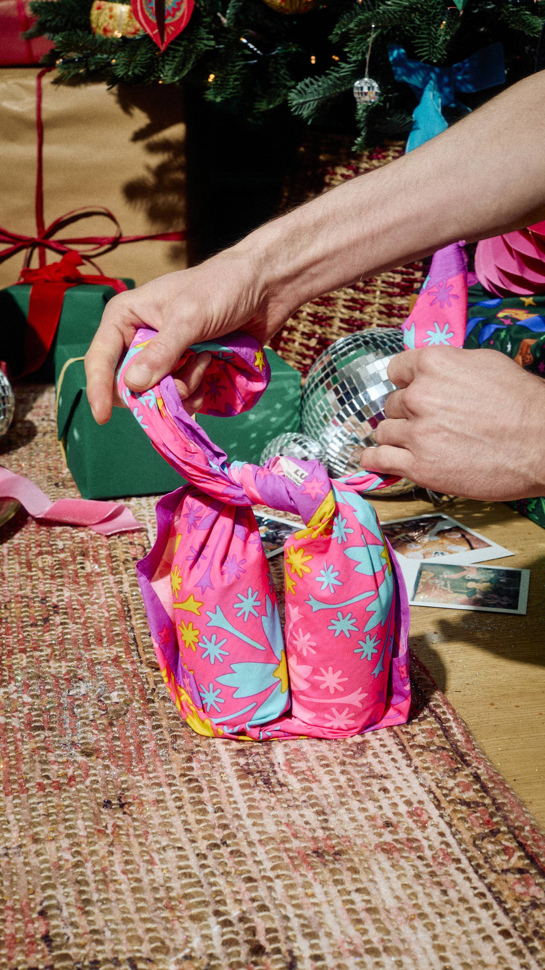 Hands tie a knot in a pink floral Lush fabric "Snow Fairy" Knot Wrap, wrapping two bottles into a gift-bag shape, set on a rug near a Christmas tree, disco balls, and instant photos.