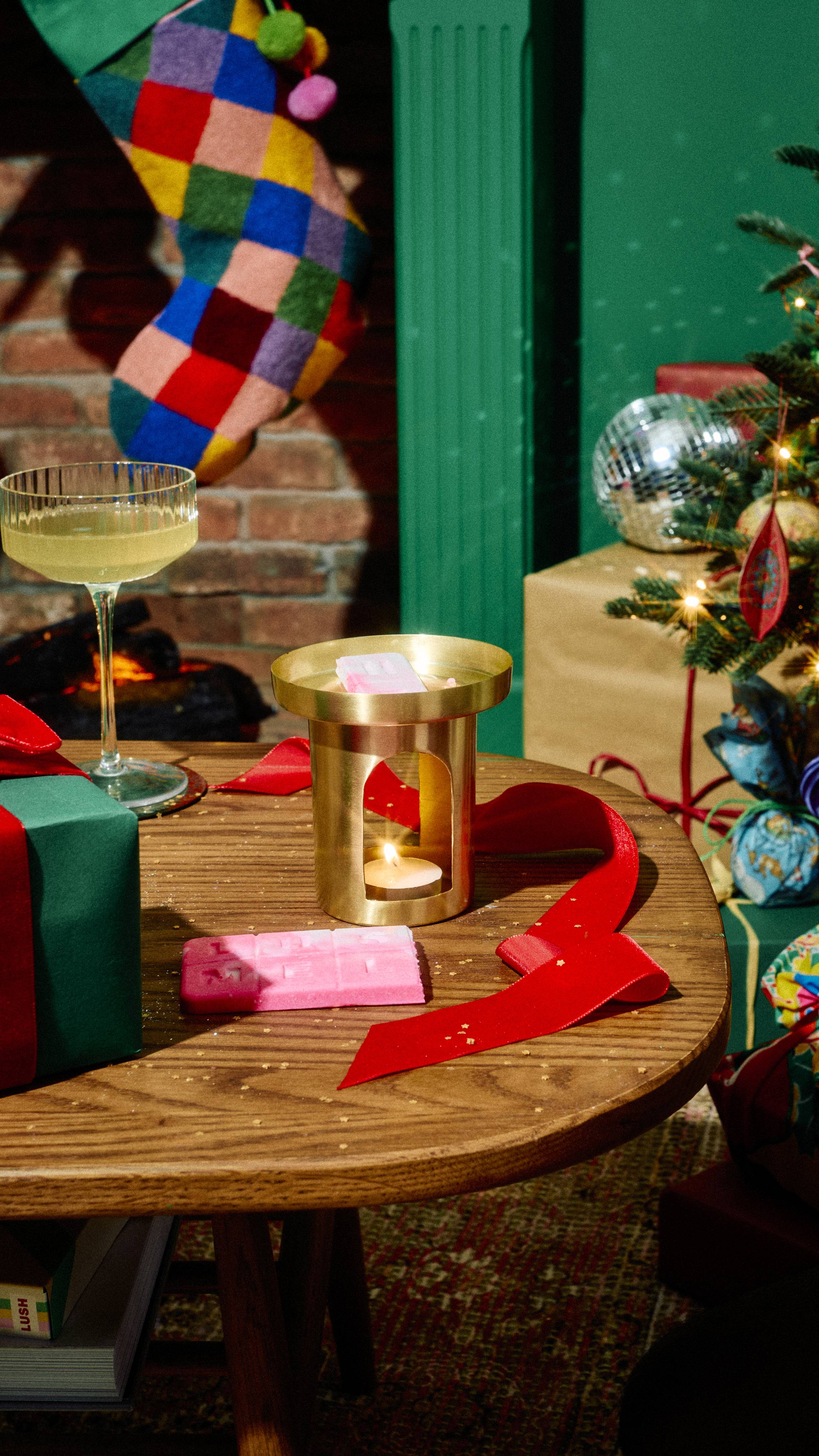 Pink Lush "Snow Fairy" wax melt pieces sit inside a brass burner with a lit tealight on a wooden table alongside a pink wax bar, cocktail glass, red ribbon, and wrapped presents in a festive room setting.