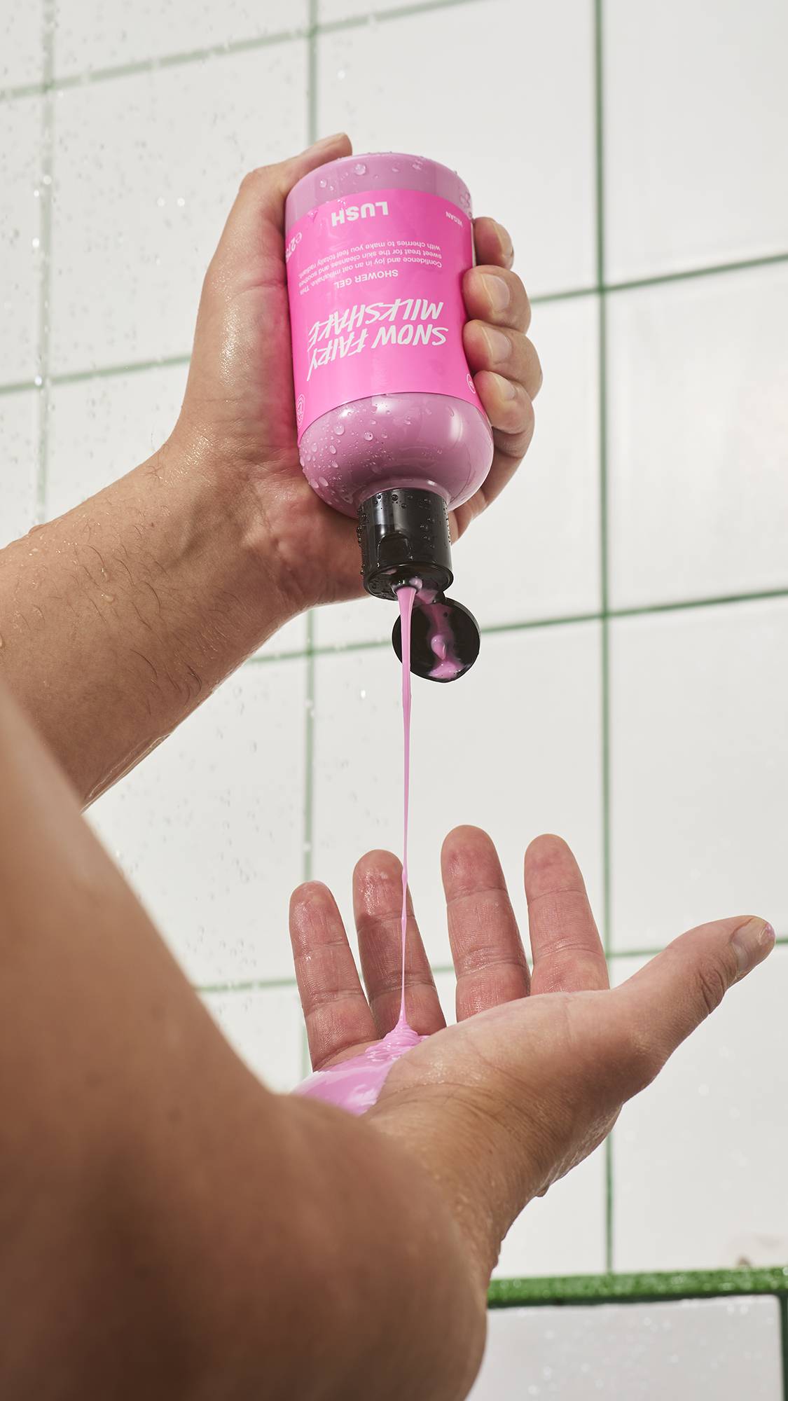 A person squeezes a bottle of Lush "Snow Fairy Milkshake" shower gel upside down, pouring a steady stream of creamy pastel pink shower gel into their open palm against a white tiled wall.