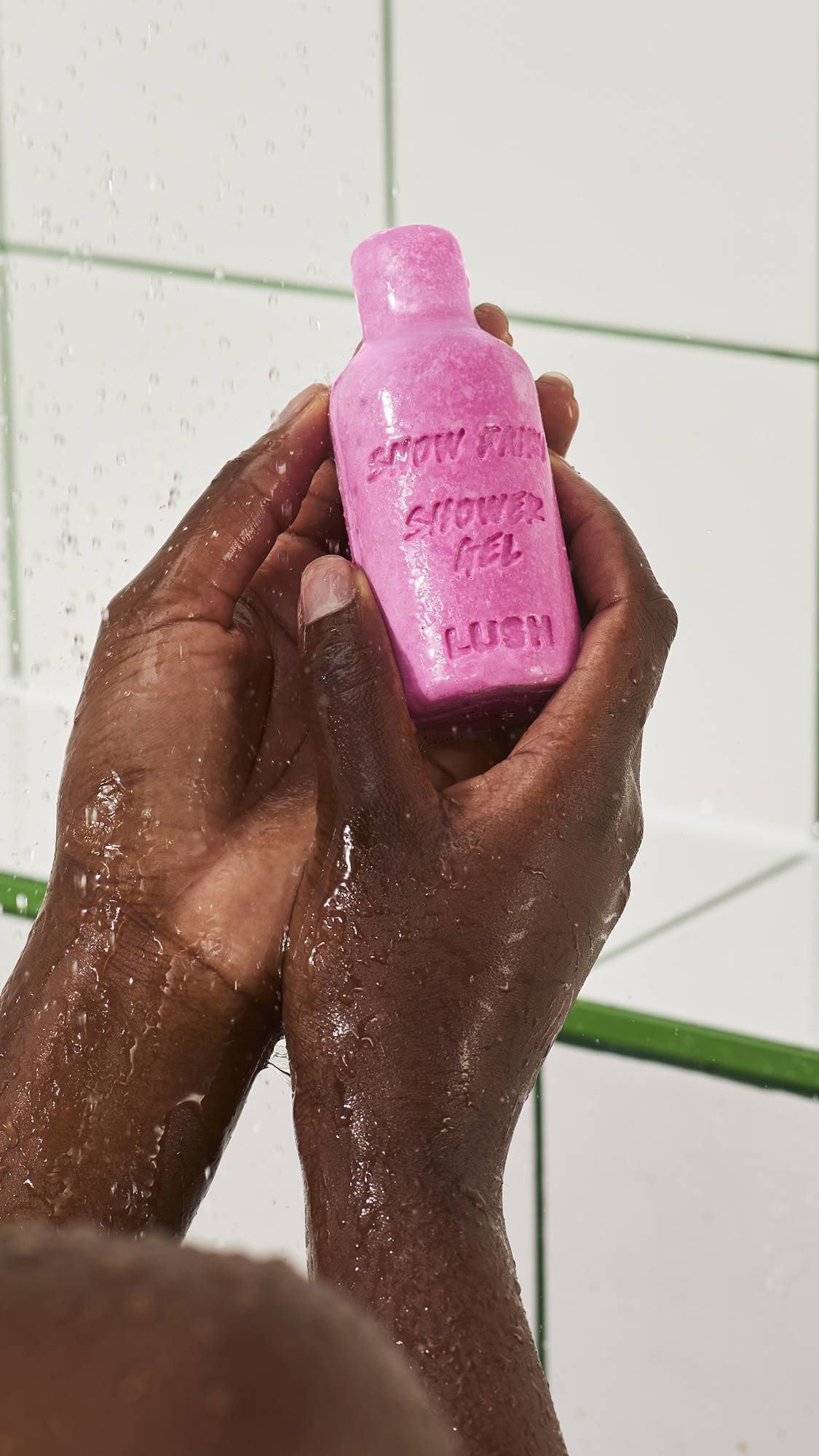A close-up view of wet hands holding a bottle-shaped bar of pink Lush Snow Fairy naked shower gel under running water, set against white tiled shower walls.