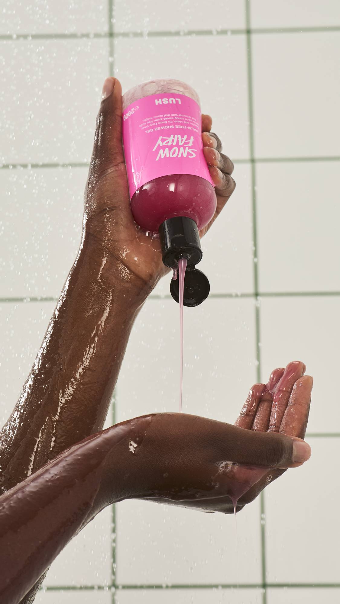 A person squeezes a bottle of Lush "Snow Fairy Palm and SLS-free" pink shower gel upside down in the shower, pouring a smooth stream of deep pink liquid into their waiting hand against a white tiled wall.