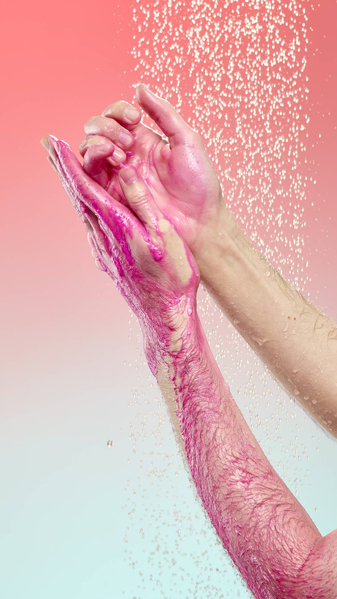 A person lathers their hands and forearm with vibrant magenta foam from Snow Fairy Pop shower powder under a spray of falling water. Set against a soft pink-to-blue gradient background.
