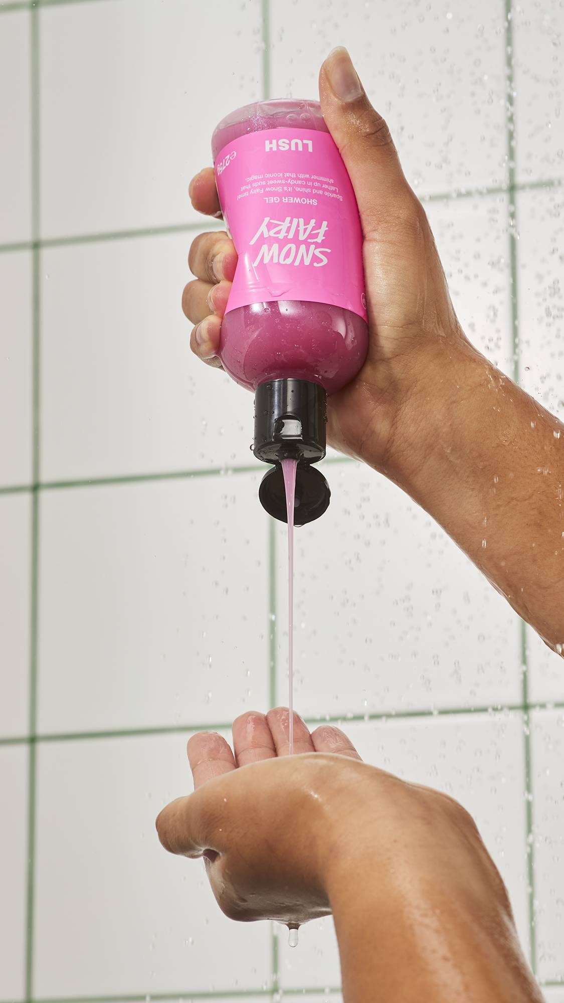 A person squeezes a bottle of Lush "Snow Fairy" pink shower gel upside down in the shower, pouring a smooth stream of shimmering pink liquid into their waiting hand against a white tiled wall.