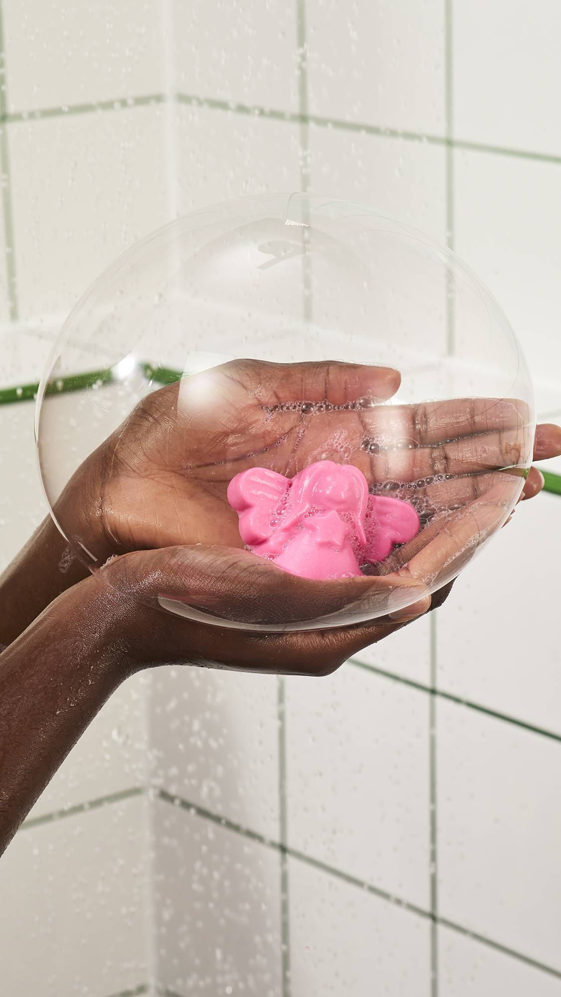 A pair of hands cupped together holds a pink Lush "Snow Fairy" shower jelly inside a clear, glowing soap bubble in the shower, against a white tiled wall background.