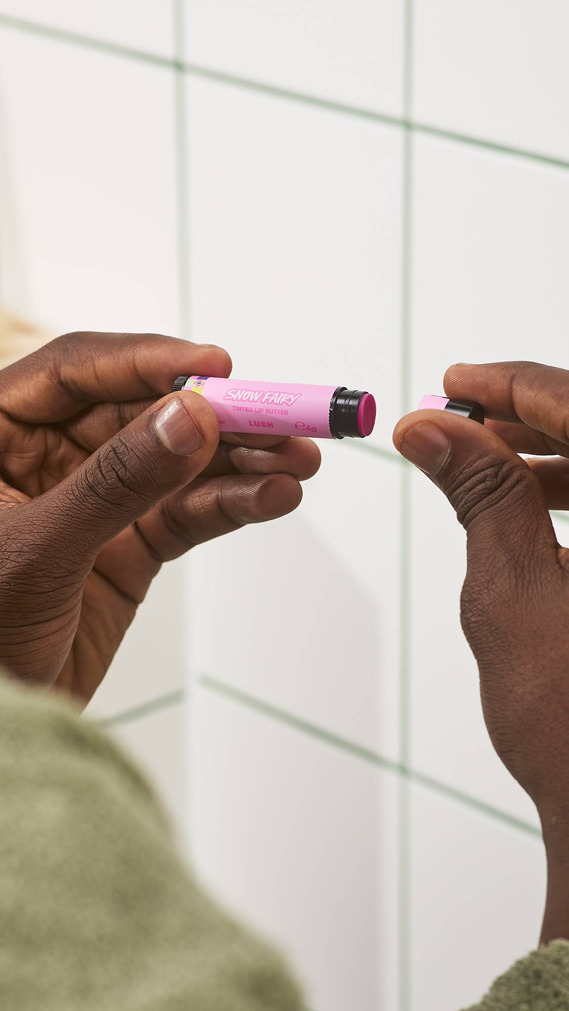 A close-up of a person's hands removing the lid from a pink tube of Lush Snow Fairy tinted lip butter to reveal the bright pink product, set against a white tiled bathroom wall background.