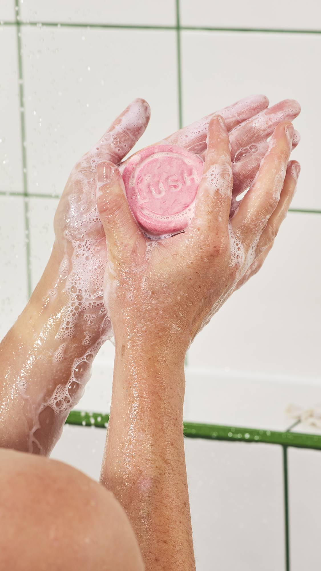 Hands hold and lather a round, pastel pink Lush s"Snow Fairy" solid shampoo bar in the shower, creating thick white suds across wet hands and arms against a white tiled wall.