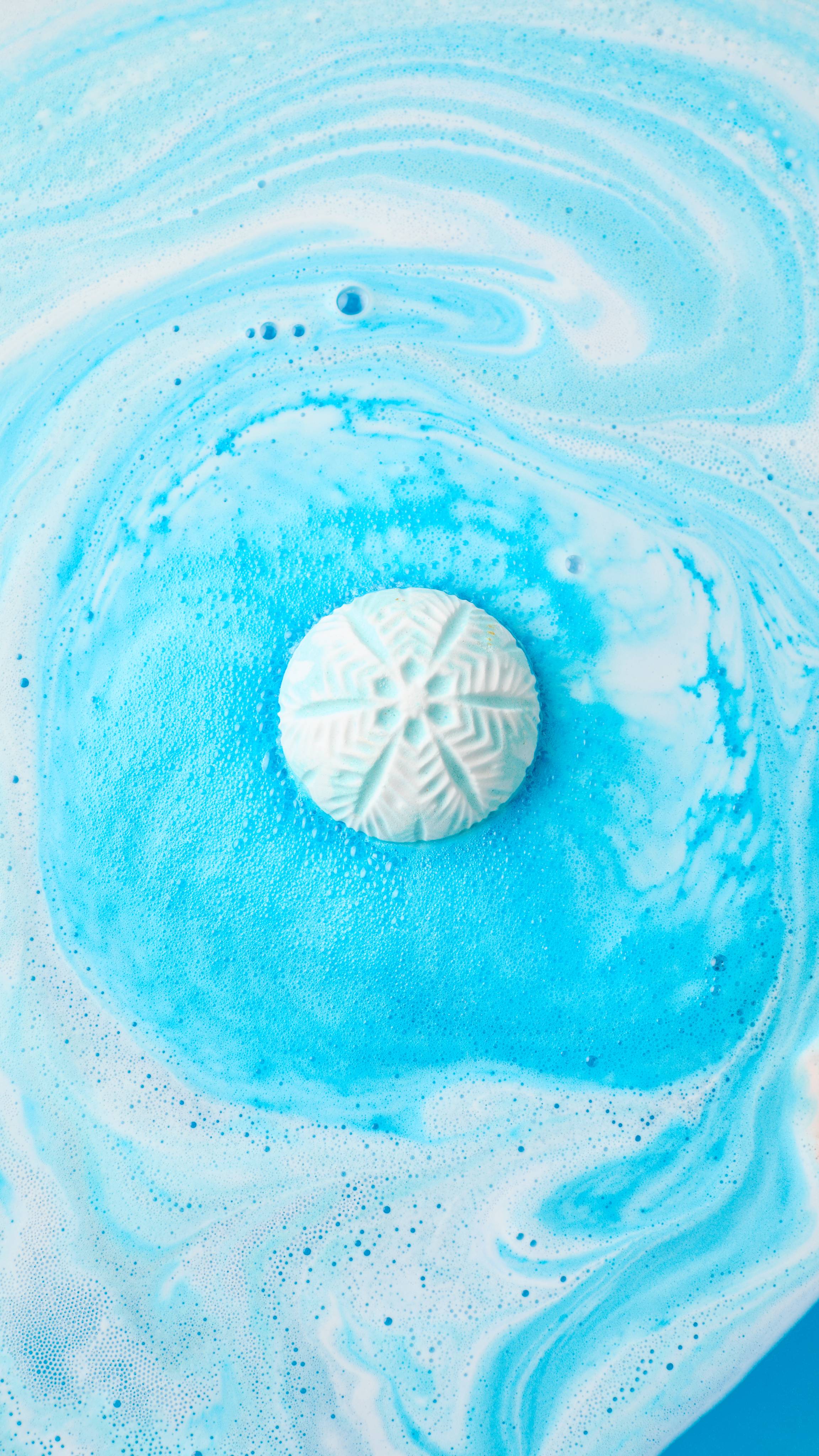 A white snowflake-embossed bath bomb dissolves at the centre of bathwater, creating a pool of vibrant sky-blue fizz encircled by swirling white foam.