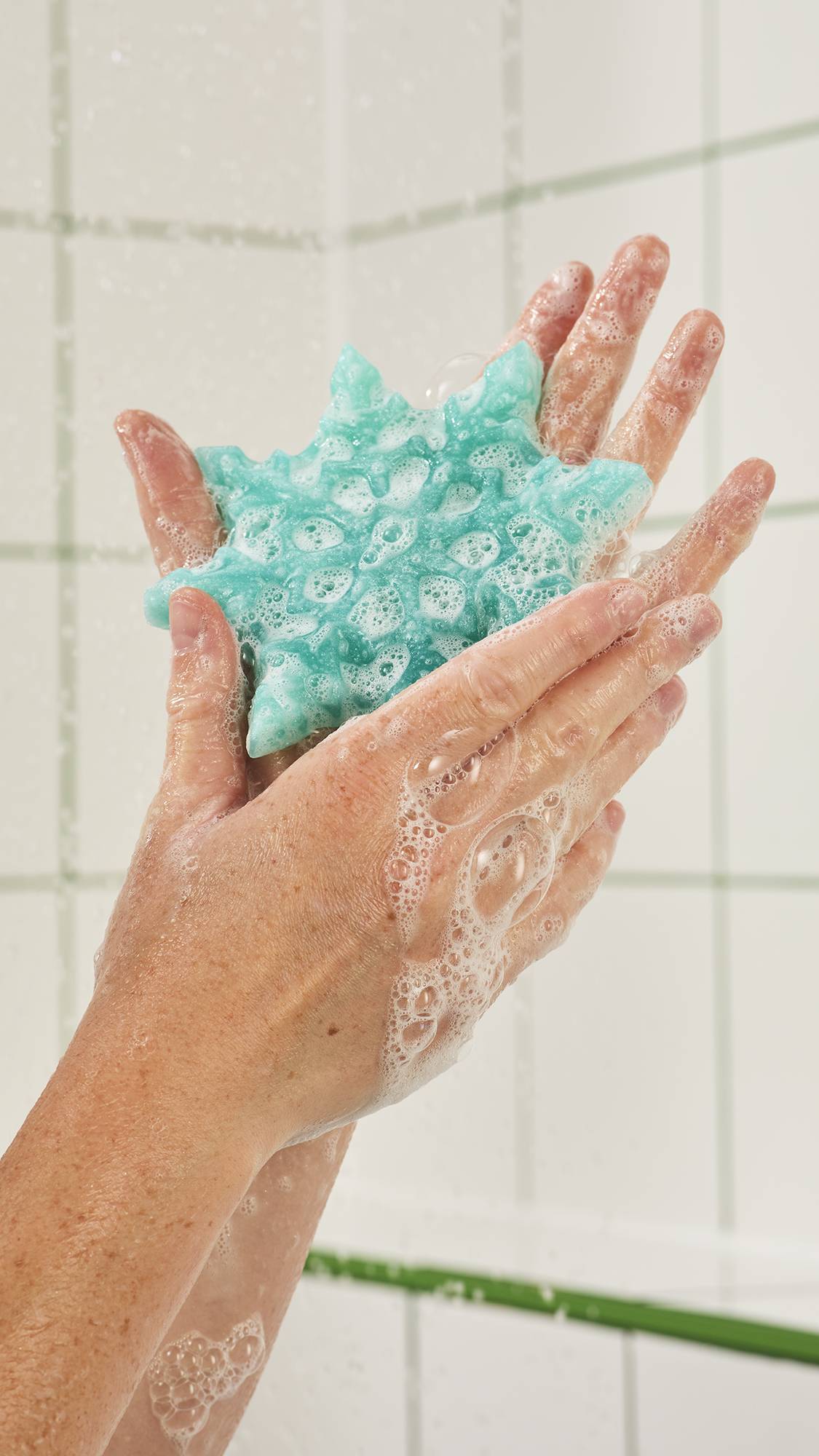 Hands lather a shimmering mint green snowflake-shaped shower gummy in the shower, creating soft white bubbles across the palms against a background of white wall tiles.