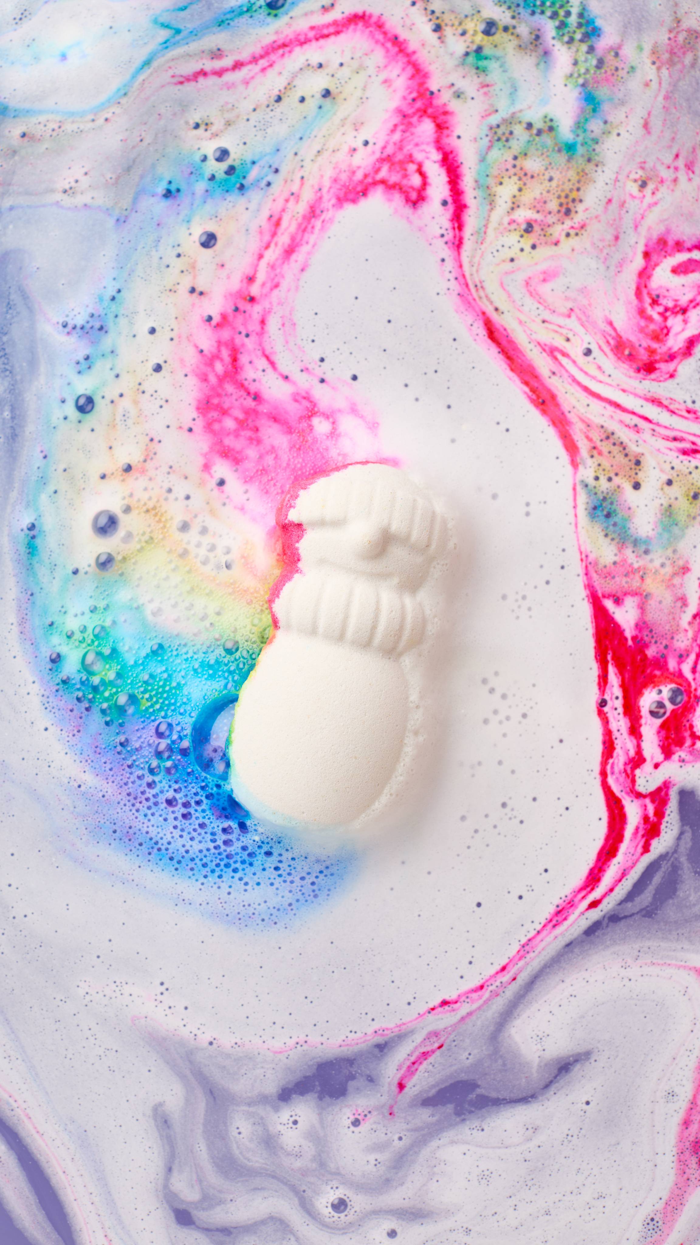 A white snowman bath bomb wearing a beanie and scarf floats on milky white bathwater, dissolving to release vibrant streaks of bright pink, cyan blue, yellow, and purple foam.