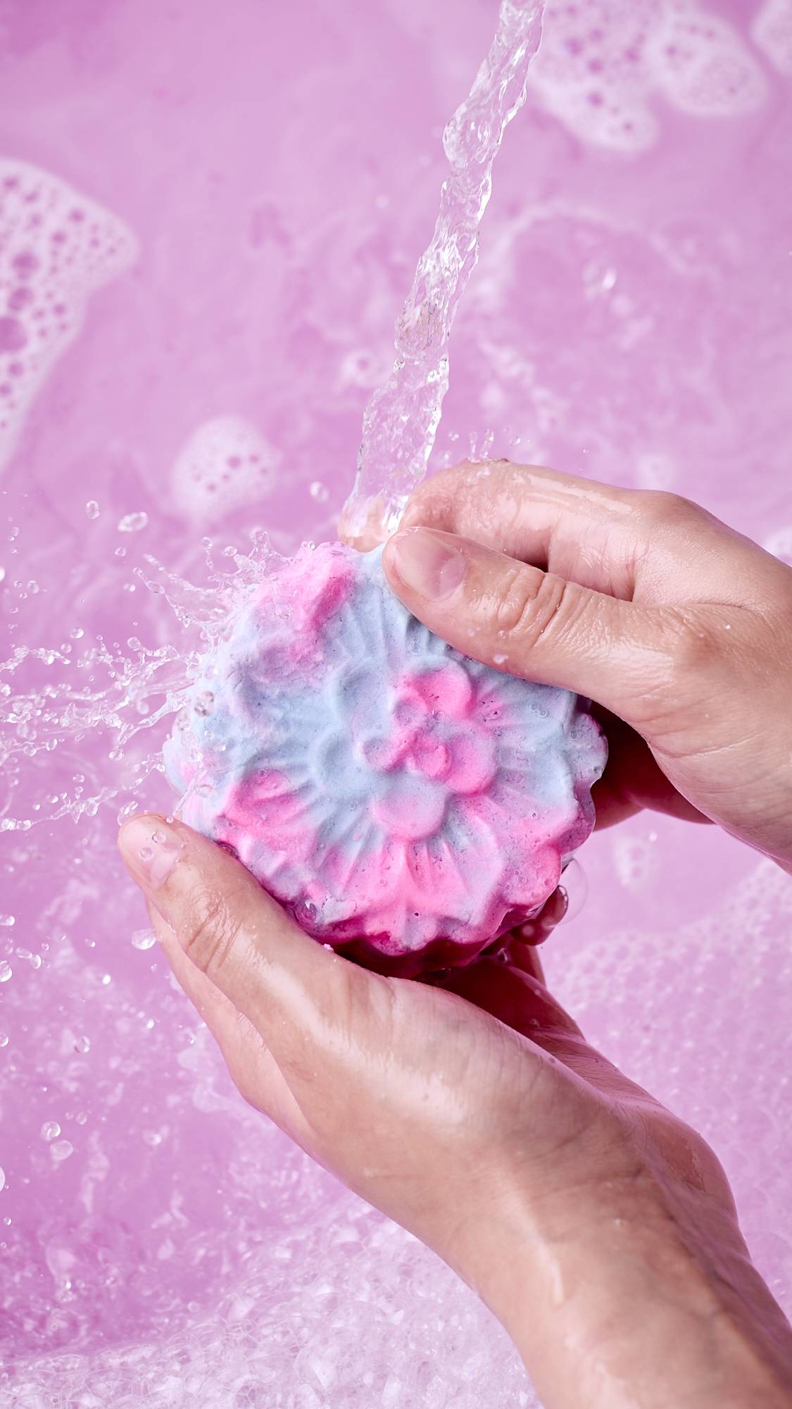 A close-up shot of hands holding a pink and blue marbled, mooncake-shaped bubble bar under a stream of running water. Water splashes off the product into a bath filled with pink, frothy foam.