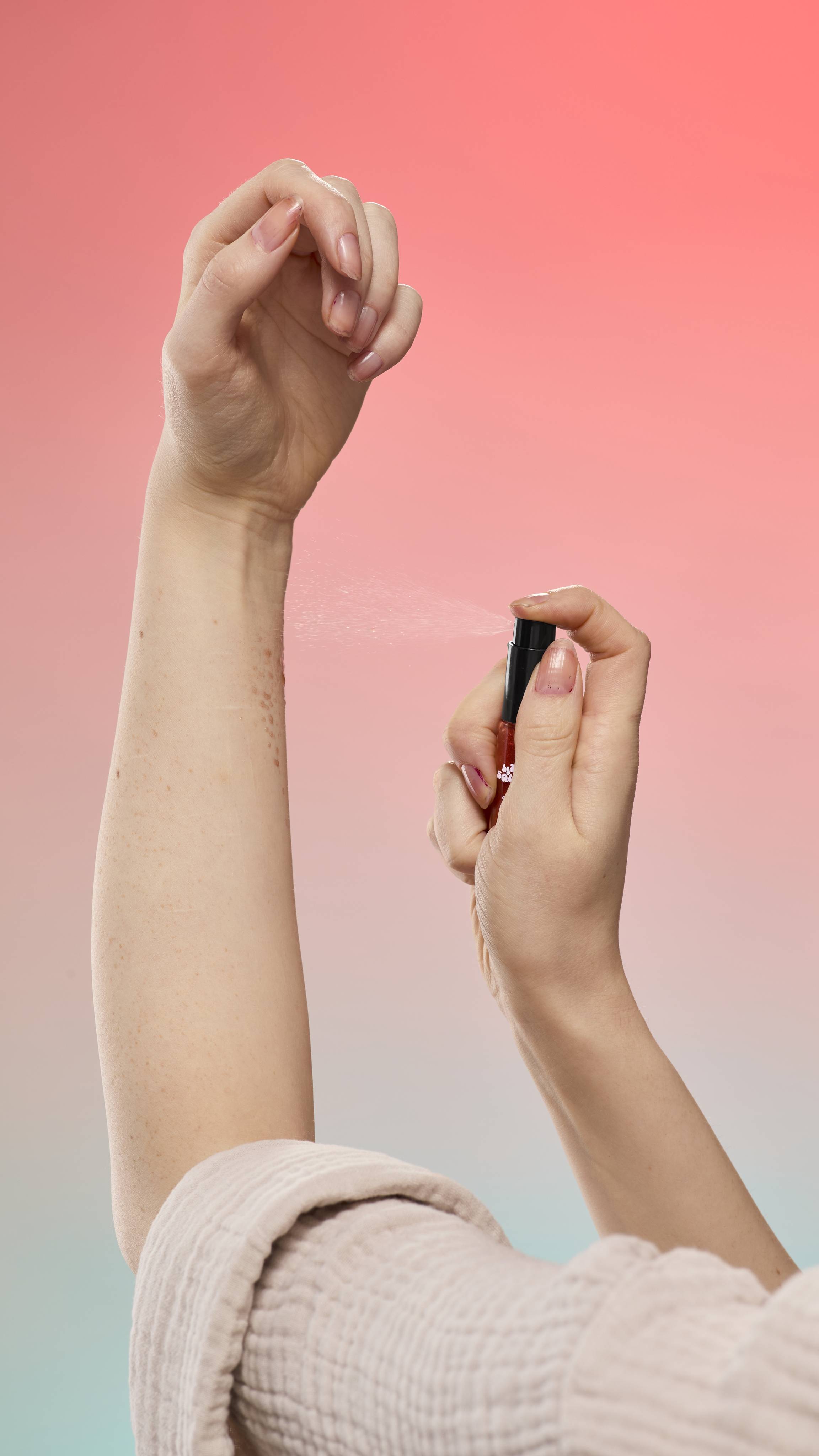 A person sprays Little Squirts Sticky Dates Mini Glitter Mist onto their arm against a peach-to-blue gradient background. They are wearing a light, textured robe while applying the fine mist from the small reddish bottle.