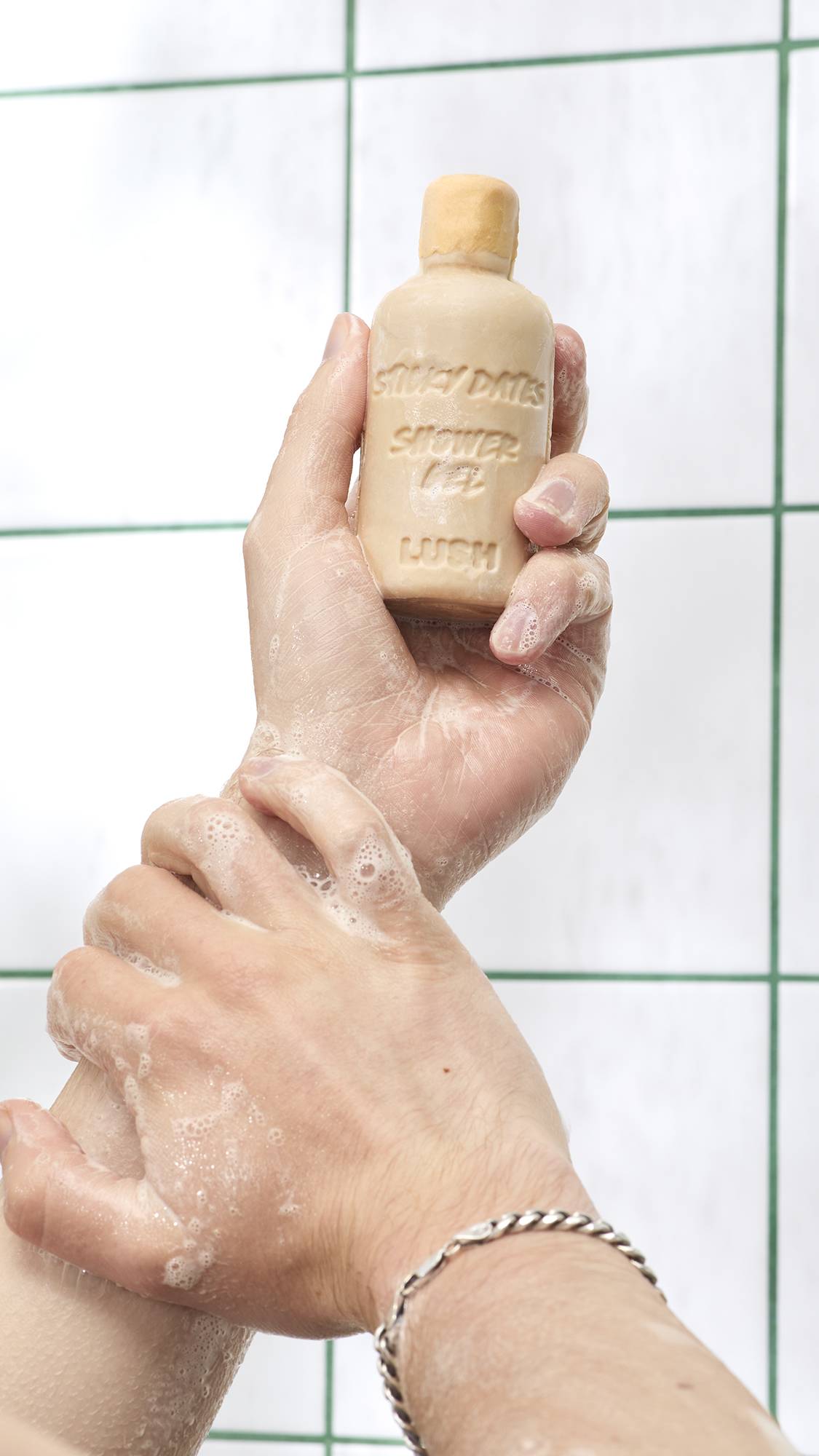 A close-up view of soapy hands holding a bottle-shaped bar of beige Lush Sticky Dates naked shower gel, washing their arm set against white tiled shower walls.