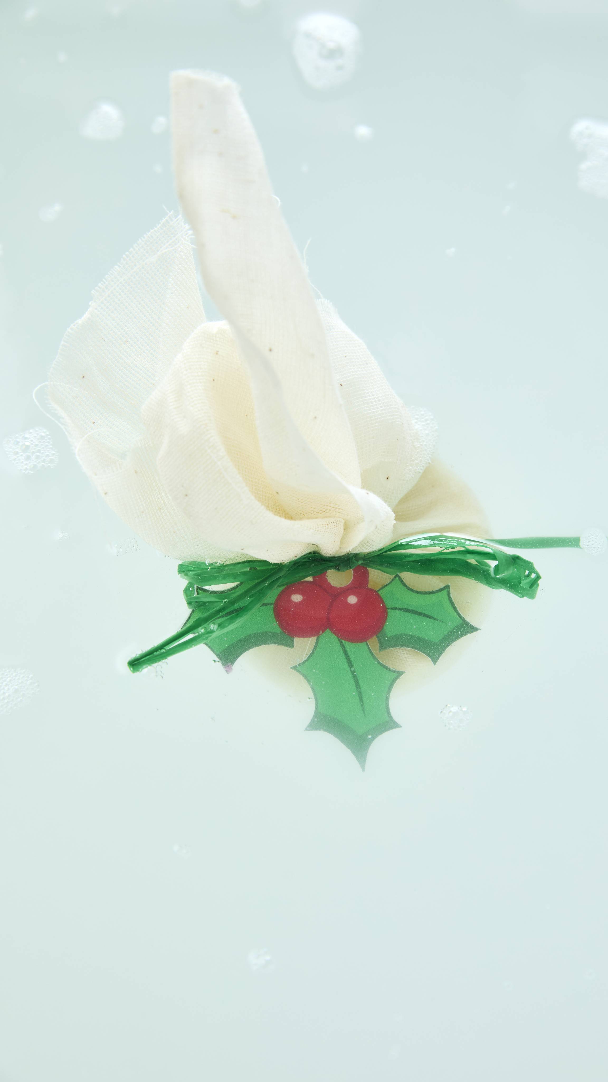 A Lush "Sticky Pudding" bath melt wrapped in cream-coloured mesh cloth with a holly tag floats in clear, lightly foamy bath water.