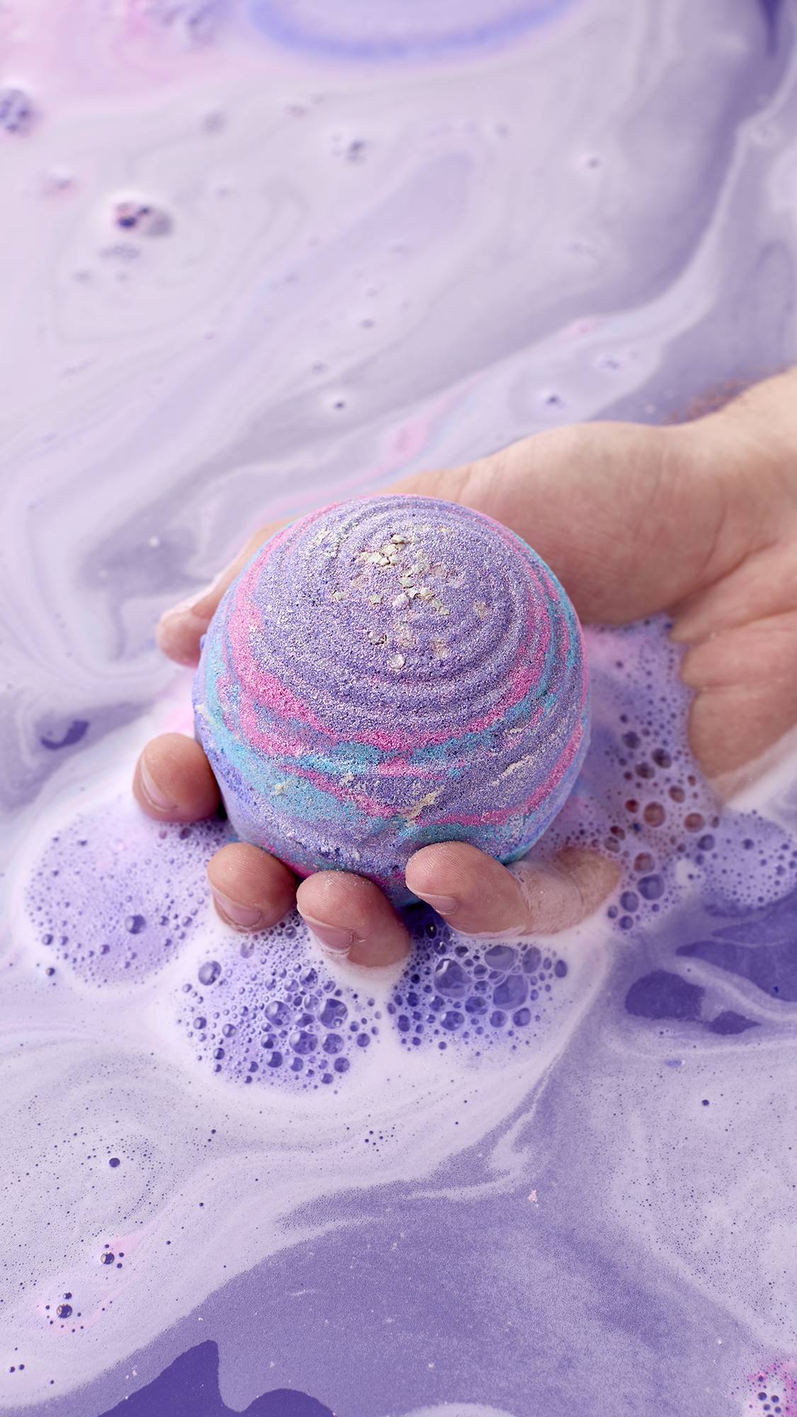 A hand holds a spherical purple, pink, and blue marbled bath bomb speckled with gold lustre, lowering it into pastel lavender bathwater as vibrant purple bubbles form around it.