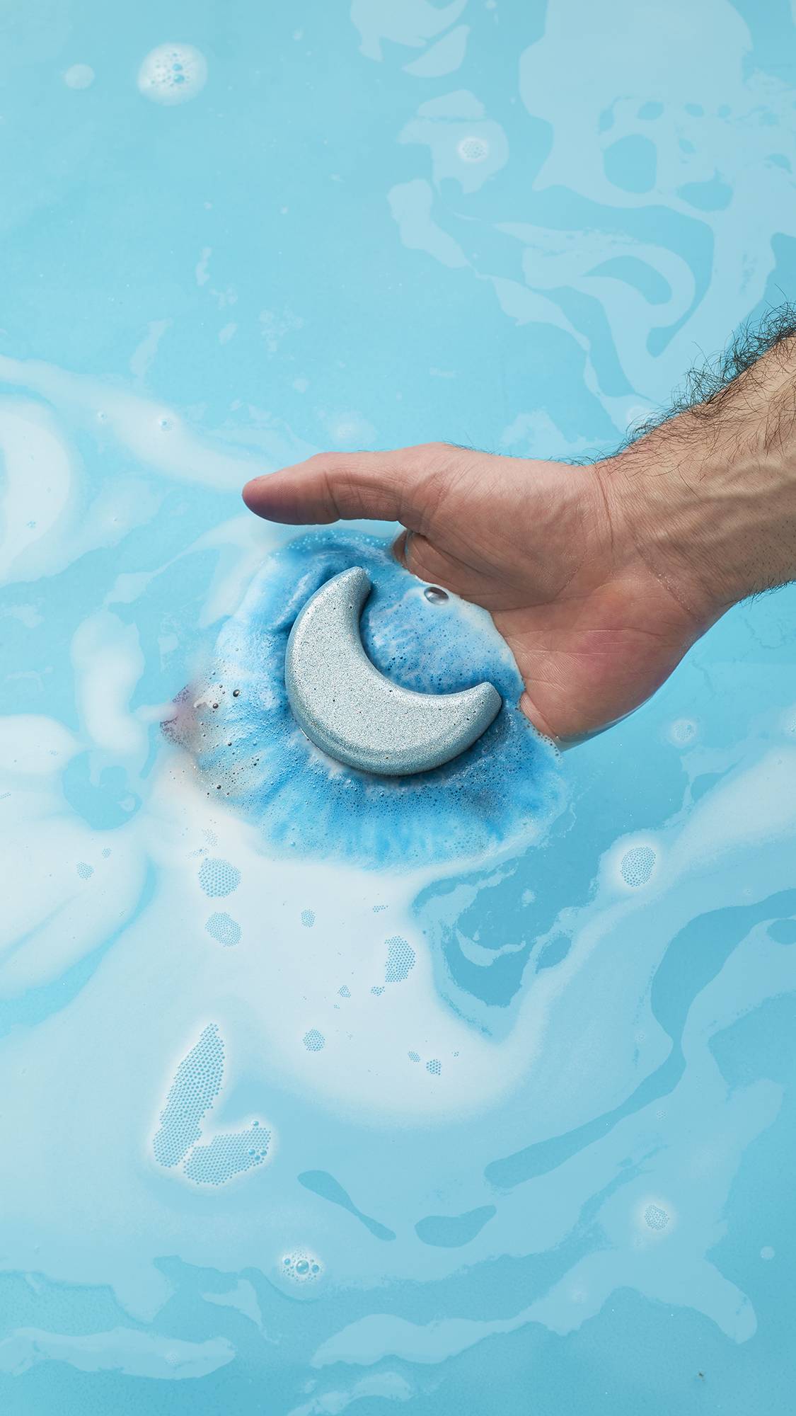 A person's hand dips the silver-blue, crescent-moon-shaped Sukoon bath bomb into bright blue water. A vibrant dark blue ring of fizz spreads out from the bath bomb as it begins to dissolve among swirls of white foam.