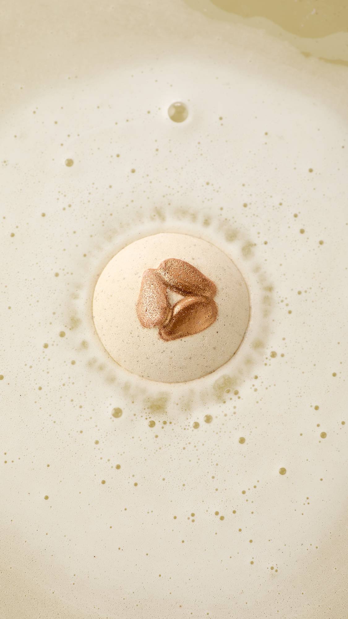 An aerial view of a speckled, off-white "Super Milk" bath bomb with 3 shimmering bronze, embossed almonds, as it begins to dissolve. It is surrounded by a ring of fine, frothy white bubbles against a creamy, pale-coloured water surface. 