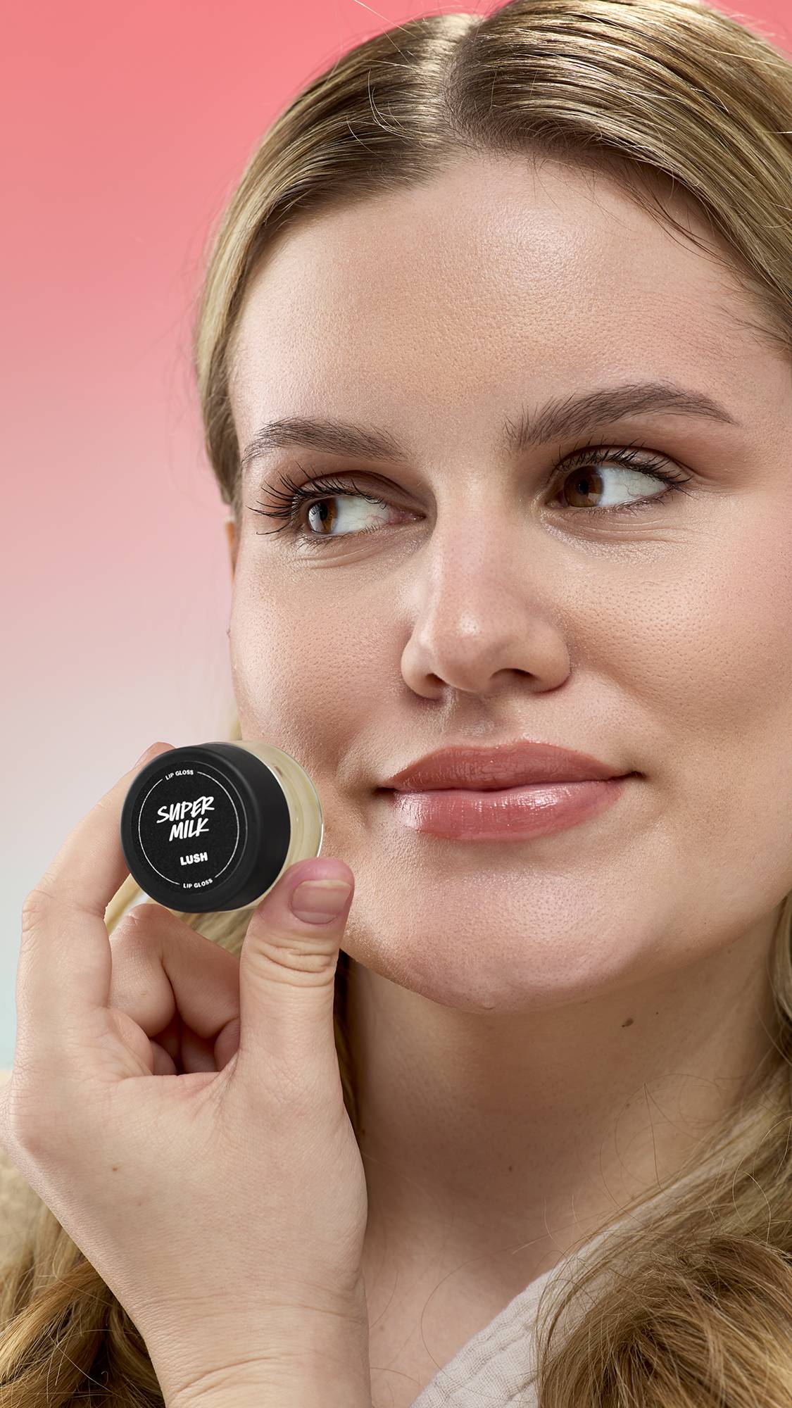 A person holds a small pot of Super Milk lip gloss by LUSH near their face. Their lips have a glossy finish. They are set against a pink gradient background.