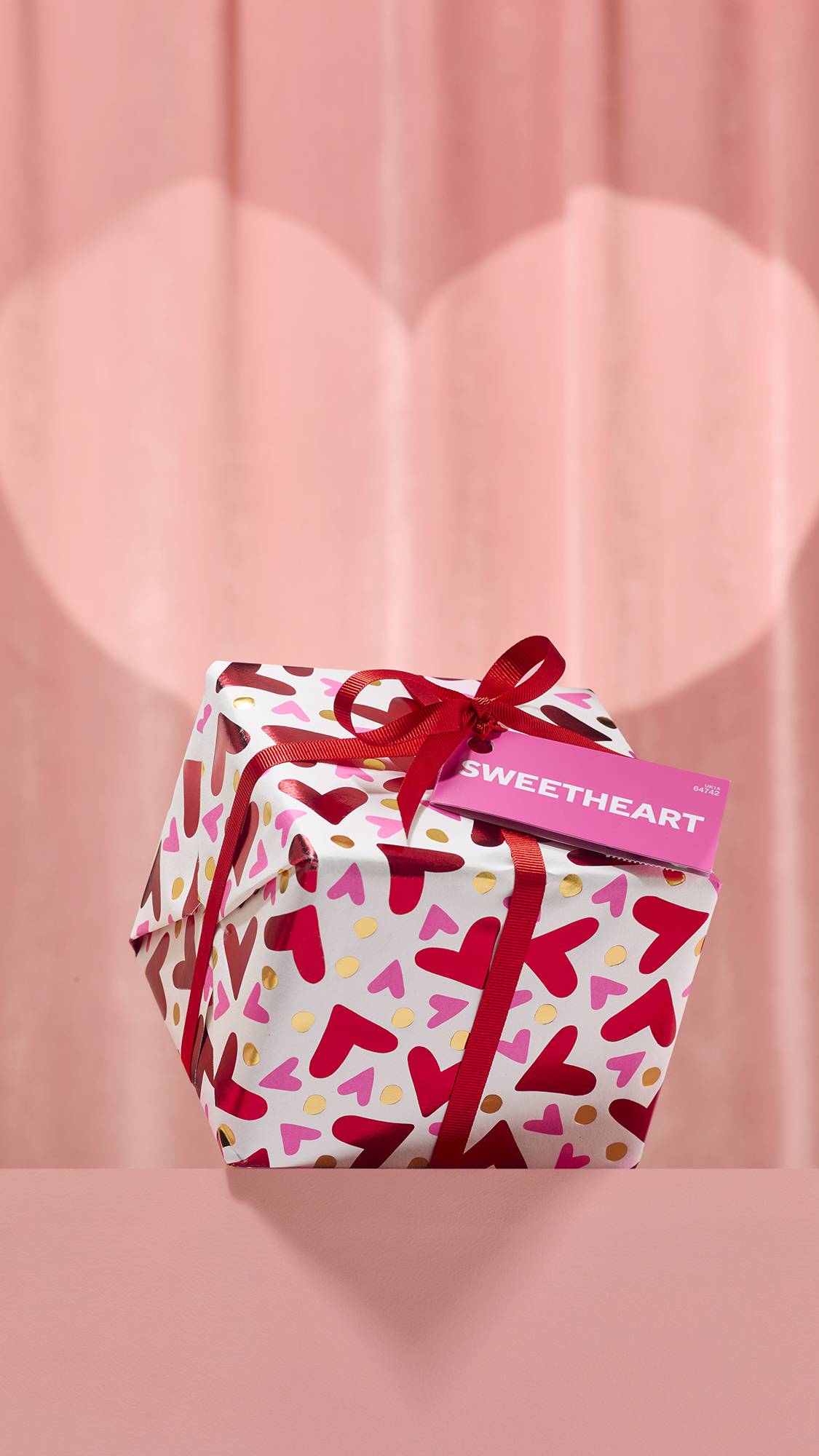 The wrapped Sweetheart Gift sits on a pink pedestal, covered in white paper with a pattern of red, pink, and metallic gold hearts. It is tied with a red ribbon and features a pink tag reading Sweetheart against a soft pink backdrop.