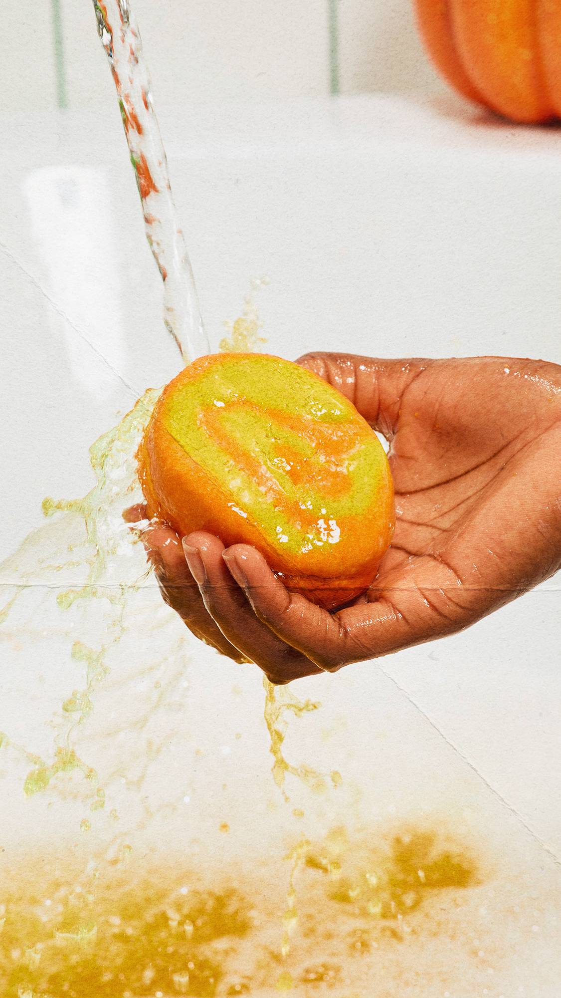 A hand holds an orange and yellow "Swirly Pumpkin" bubble bar under running tap water in a white bath, splashing and creating warm, golden-toned water below.