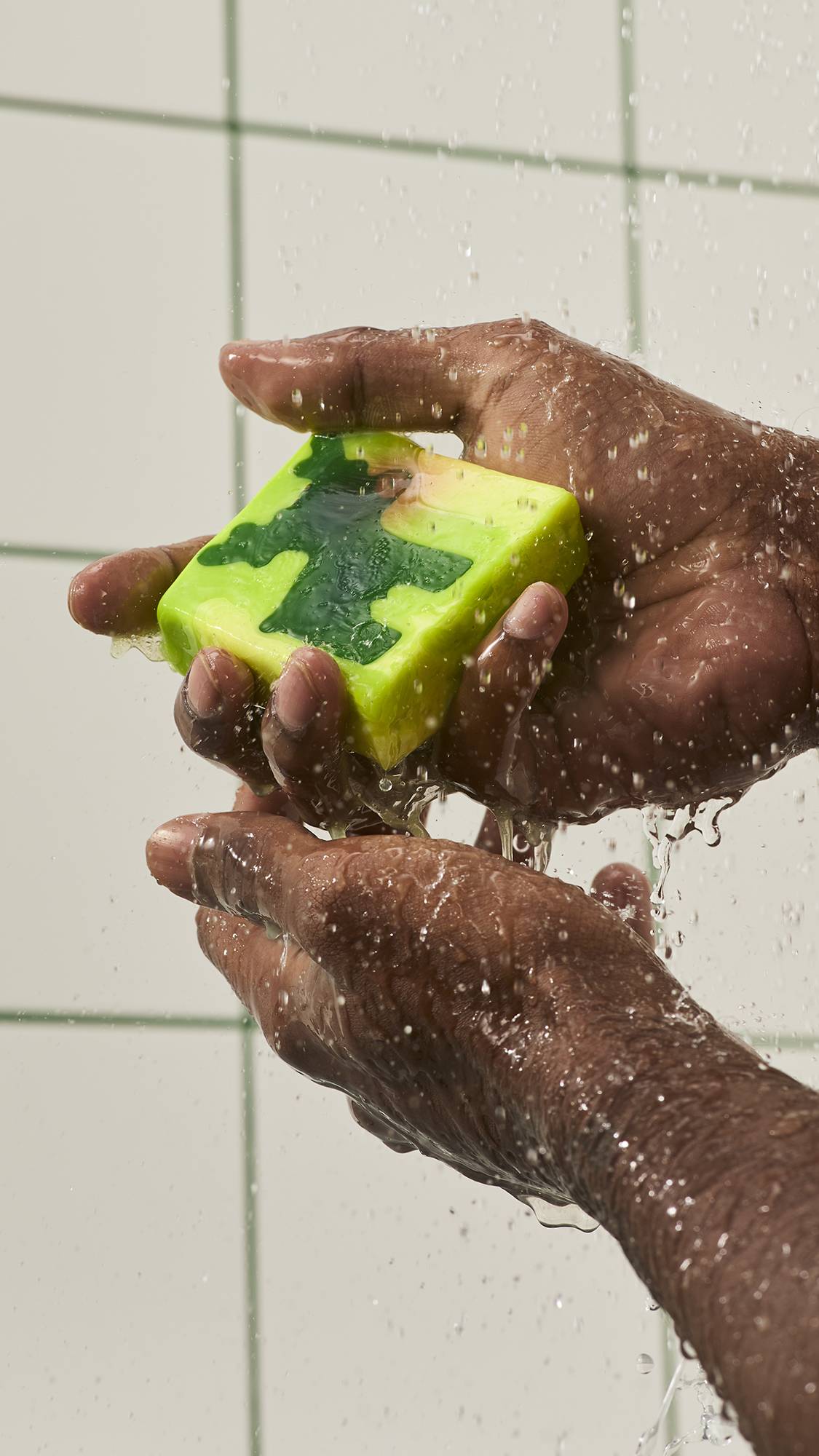 Hands hold and turn a square lime green soap featuring a dark green reindeer motif under streaming shower water against a white tiled wall background.
