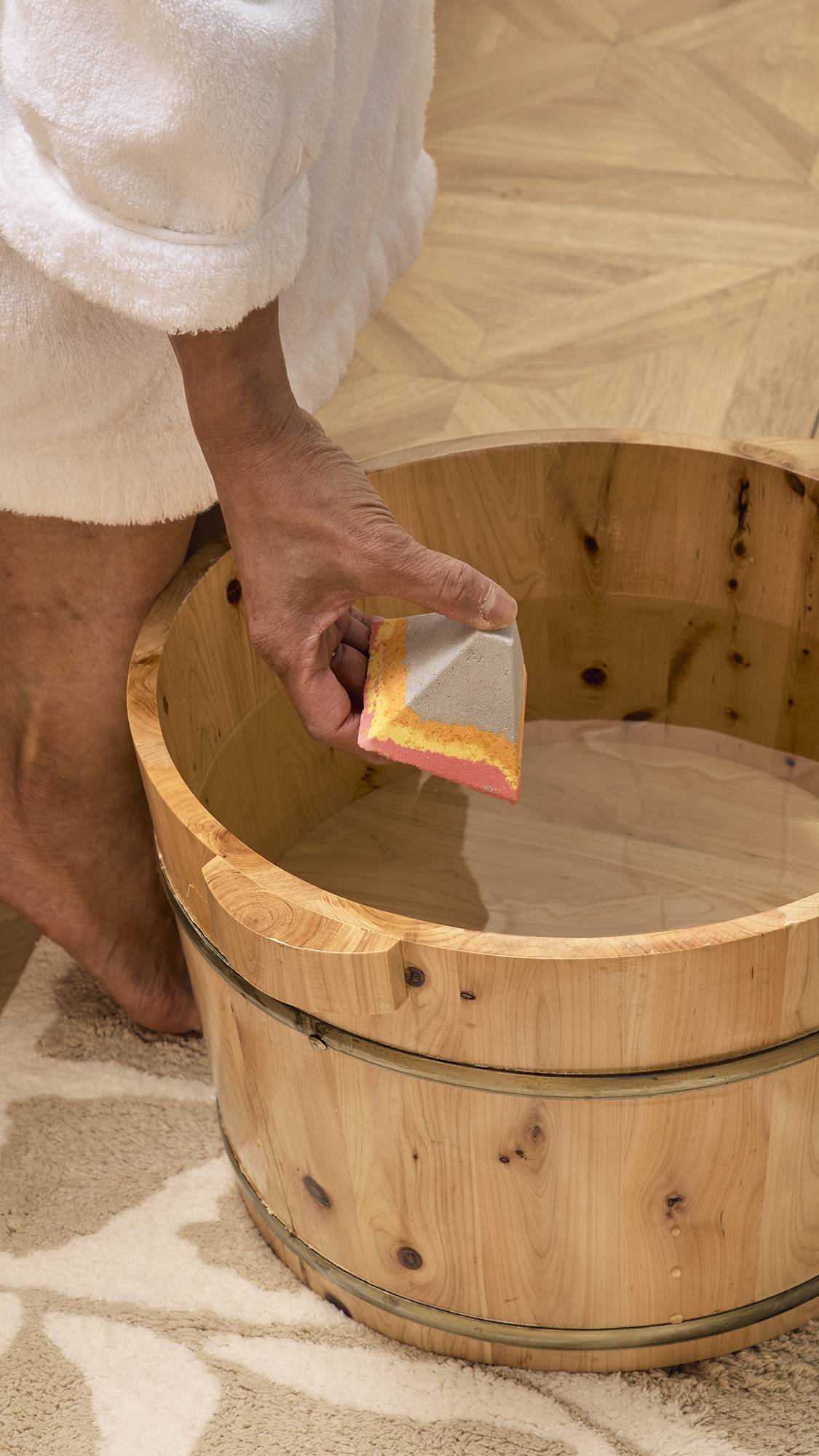 A model wearing a white dressing gown dips a pyramid-shaped "Volcano" foot scrub into a wooden half-barrel tub. The grey, orange, and pink bath bomb is held just above the clear water, ready to be dissolved for a relaxing foot spa. 