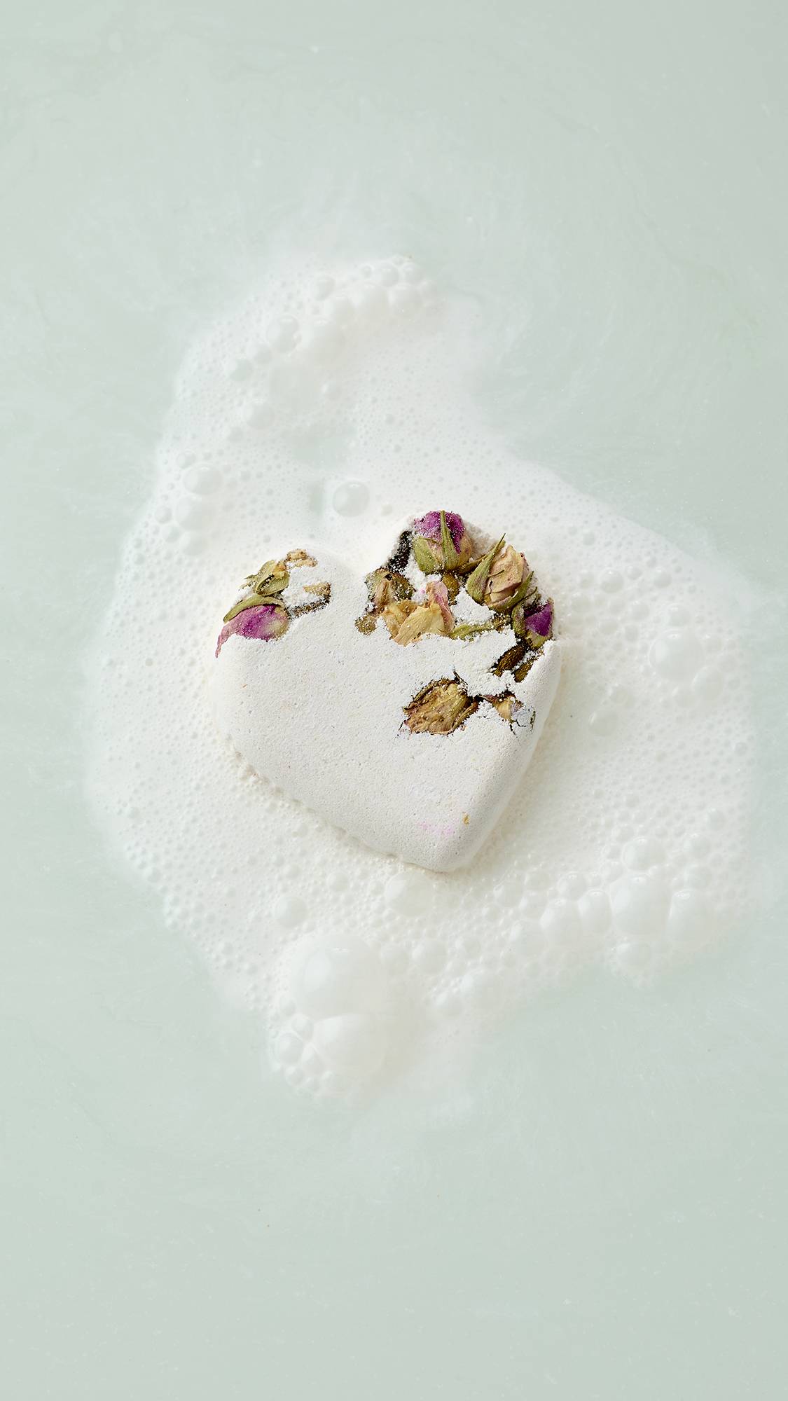 The image shows an aerial view of a white, heart-shaped Tisty Tosty bath bomb decorated with dried rosebuds floating in pale water. It is surrounded by a patch of white, frothy bubbles as it begins to dissolve.