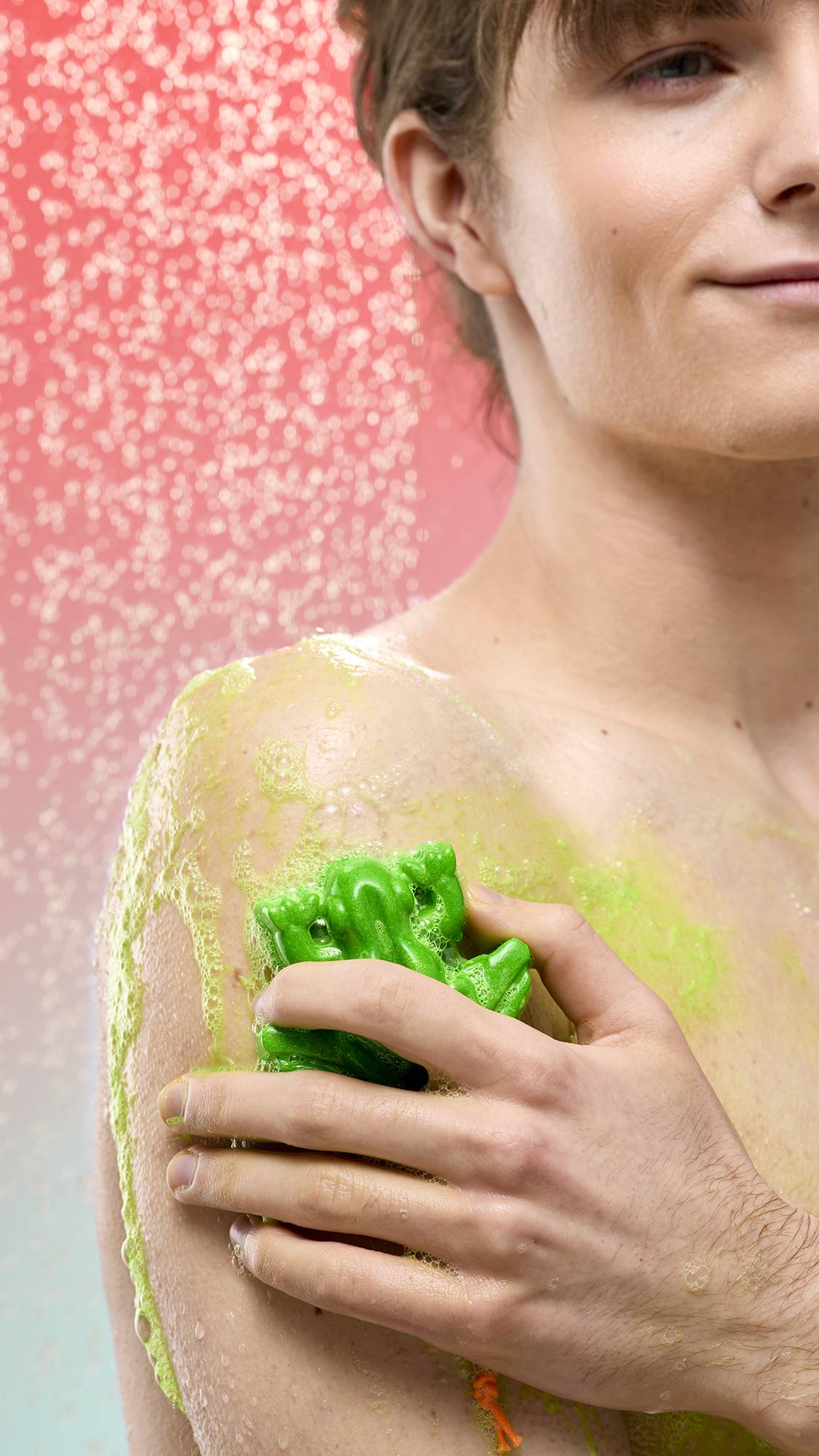 A person lathers their shoulder with the bright green, frog-shaped Tree Hugger soap under a spray of falling water. Vibrant lime-green foam covers their skin against a soft pink-to-blue gradient background.