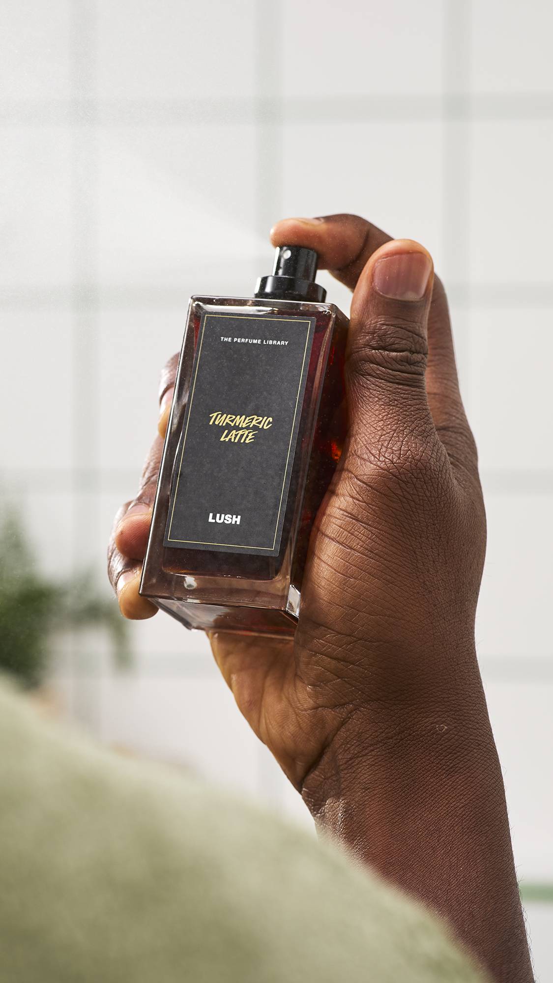 A close-up of a hand holding a glass bottle of Lush Turmeric Latte perfume, with a finger pressing down on the black spray nozzle to release a fine mist, set against a white tiled bathroom background.
