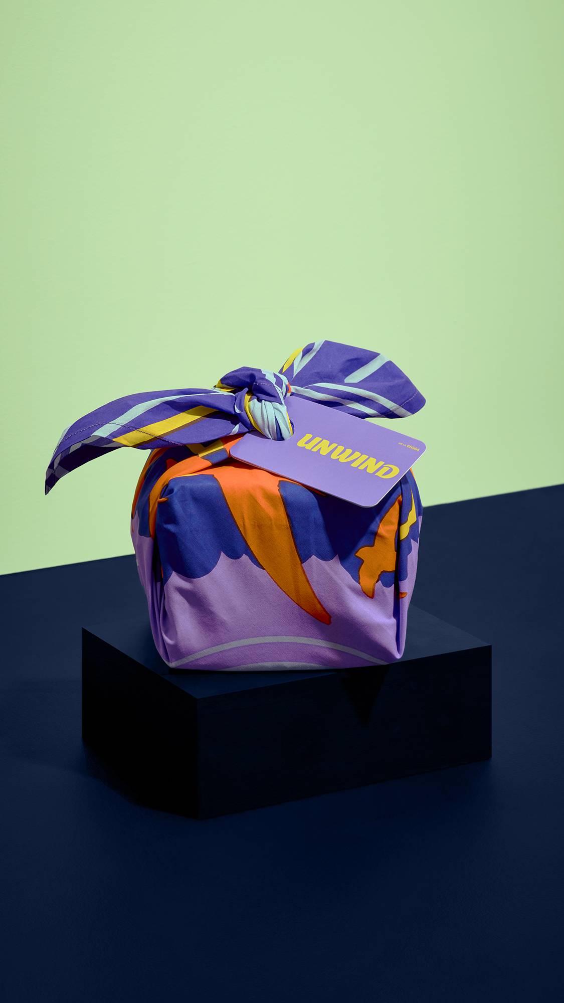 A gift wrapped in vibrant purple and orange pattern fabric sits on a dark blue pedestal against a light green wall, tagged with a lilac label reading "UNWIND" in yellow text.