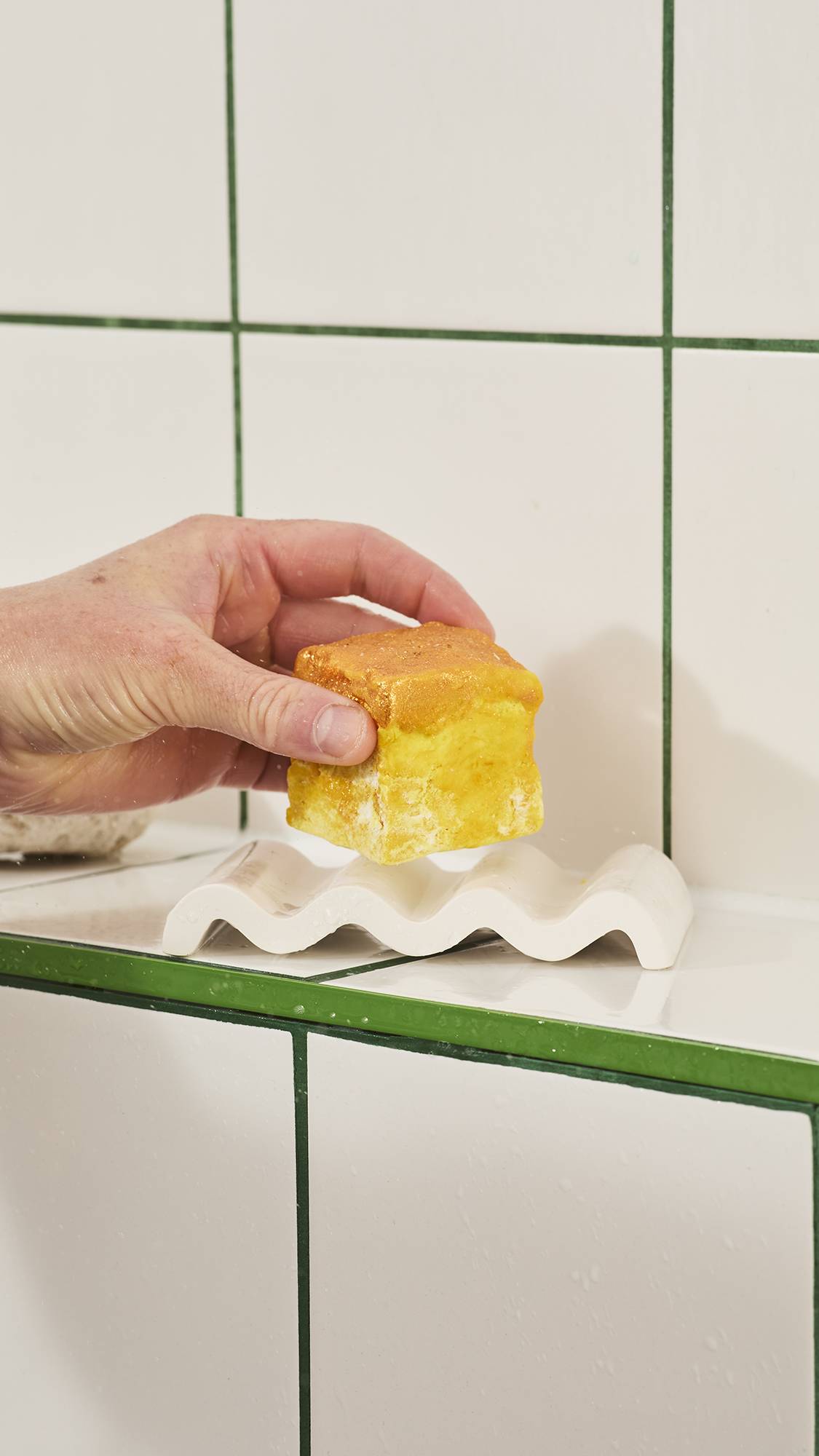 A hand lifts a small, textured yellow cube of Lush "Vanilla Delight" soap off a wavy white ceramic soap dish resting on a tiled shelf against a white tiled wall background.