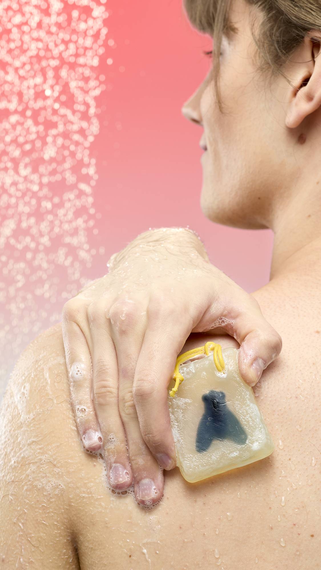 A person lathers their shoulder with Waiter! There's A Fly In My Soap under falling water. The translucent yellow bar contains a dark fly silhouette and a yellow cord. Set against a pink-to-blue gradient background.
