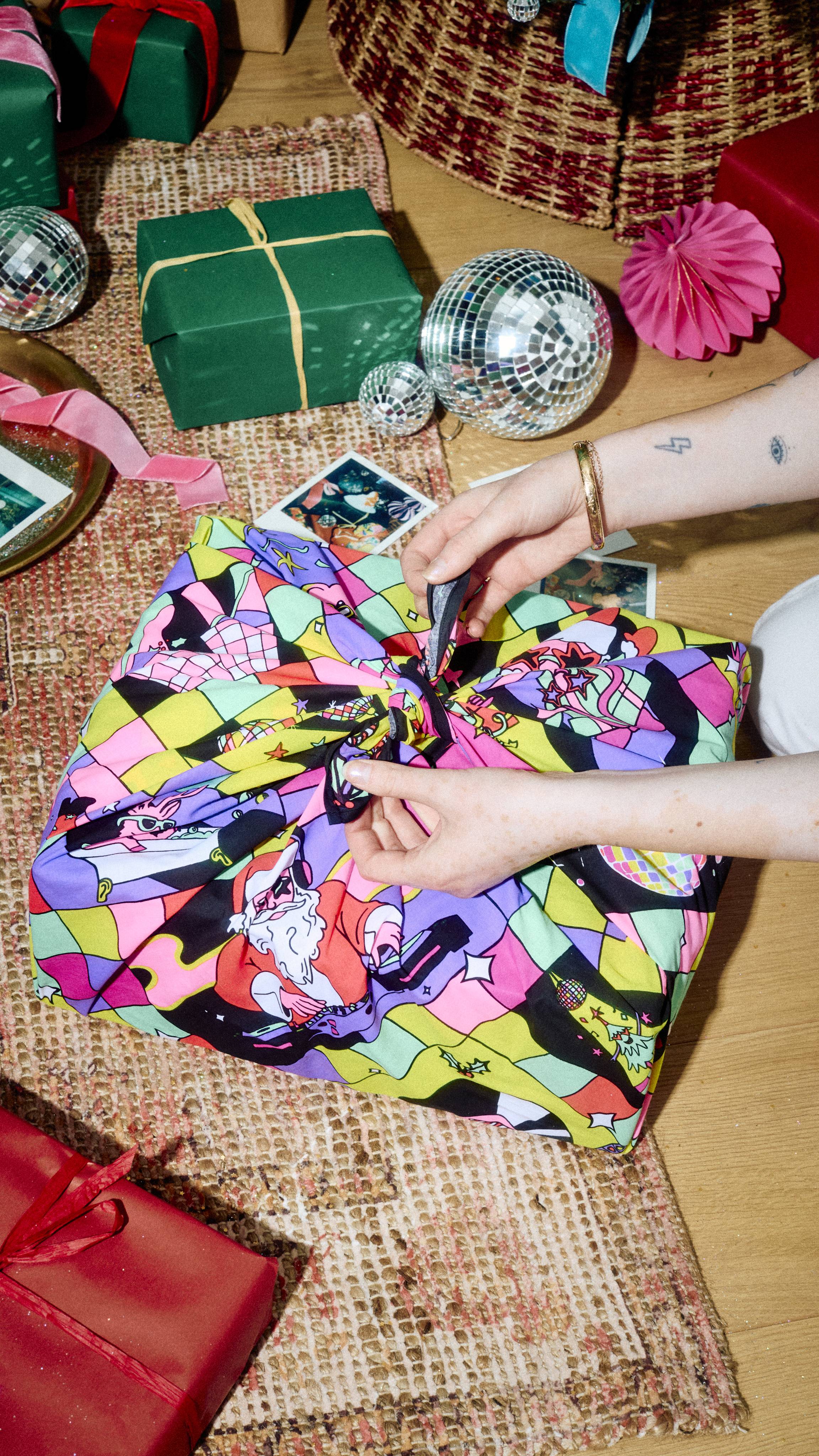 Hands tie a knot on a colourful, festive disco-patterned Lush fabric Knot Wrap covering a gift, set on a rug alongside wrapped presents, disco balls, and instant photos.