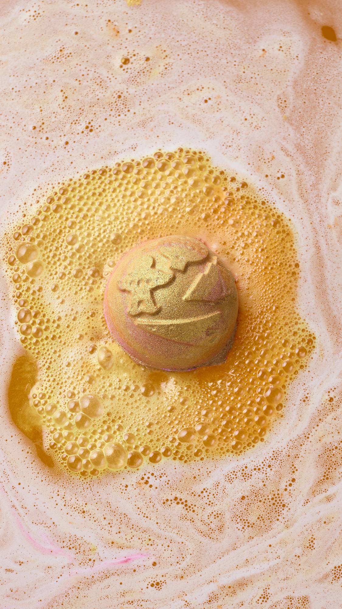 A golden, Lush "Yog Nog" bath bomb dissolves in bathwater, surrounded by a pool of rich, golden-yellow foam encircled by light peach and white froth.