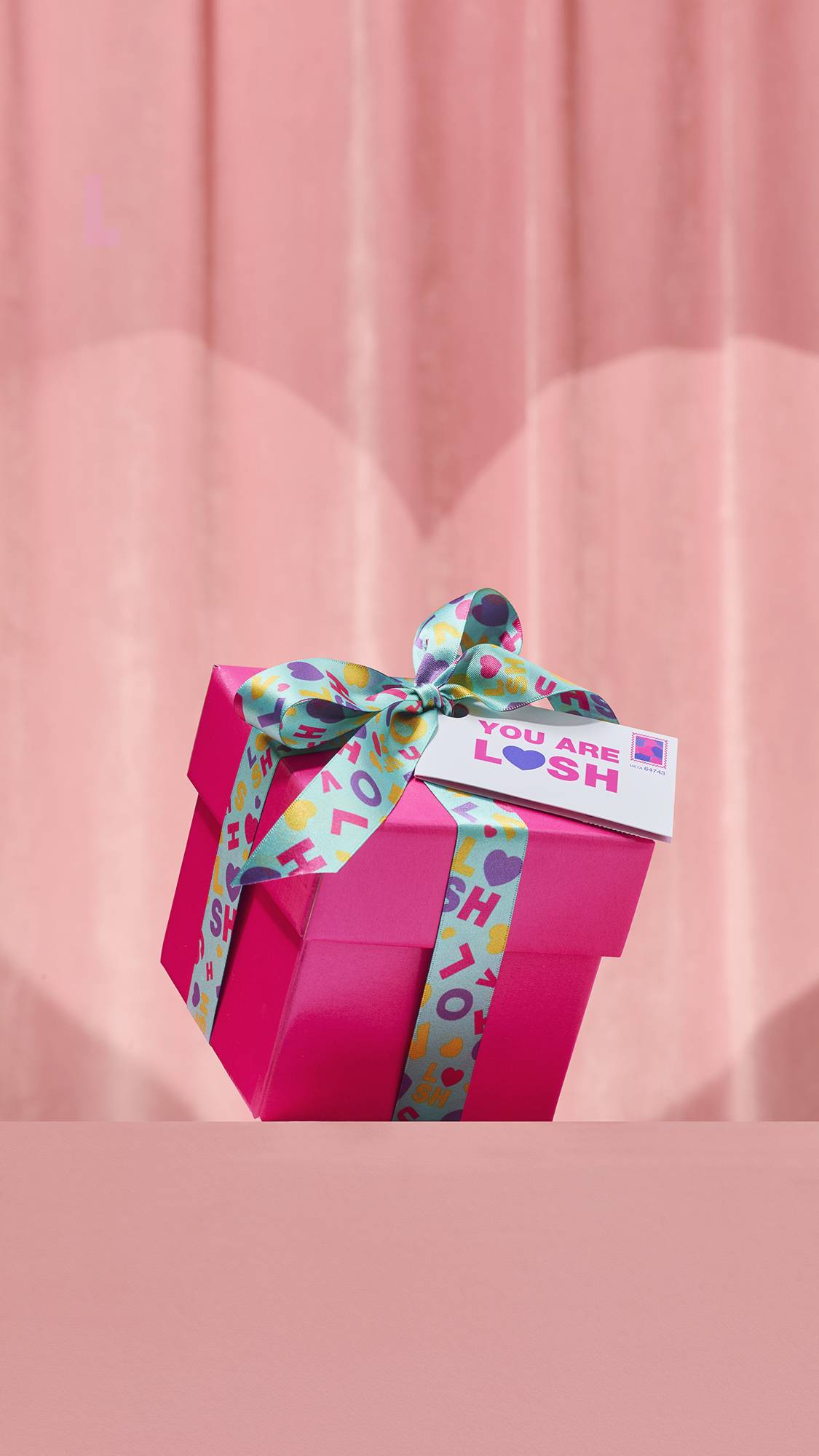 The hot pink You Are Lush Gift box is tied with a teal ribbon featuring colorful text and hearts. A tag attached to the bow reads You Are Lush with a purple heart, set against a soft pink curtain background.