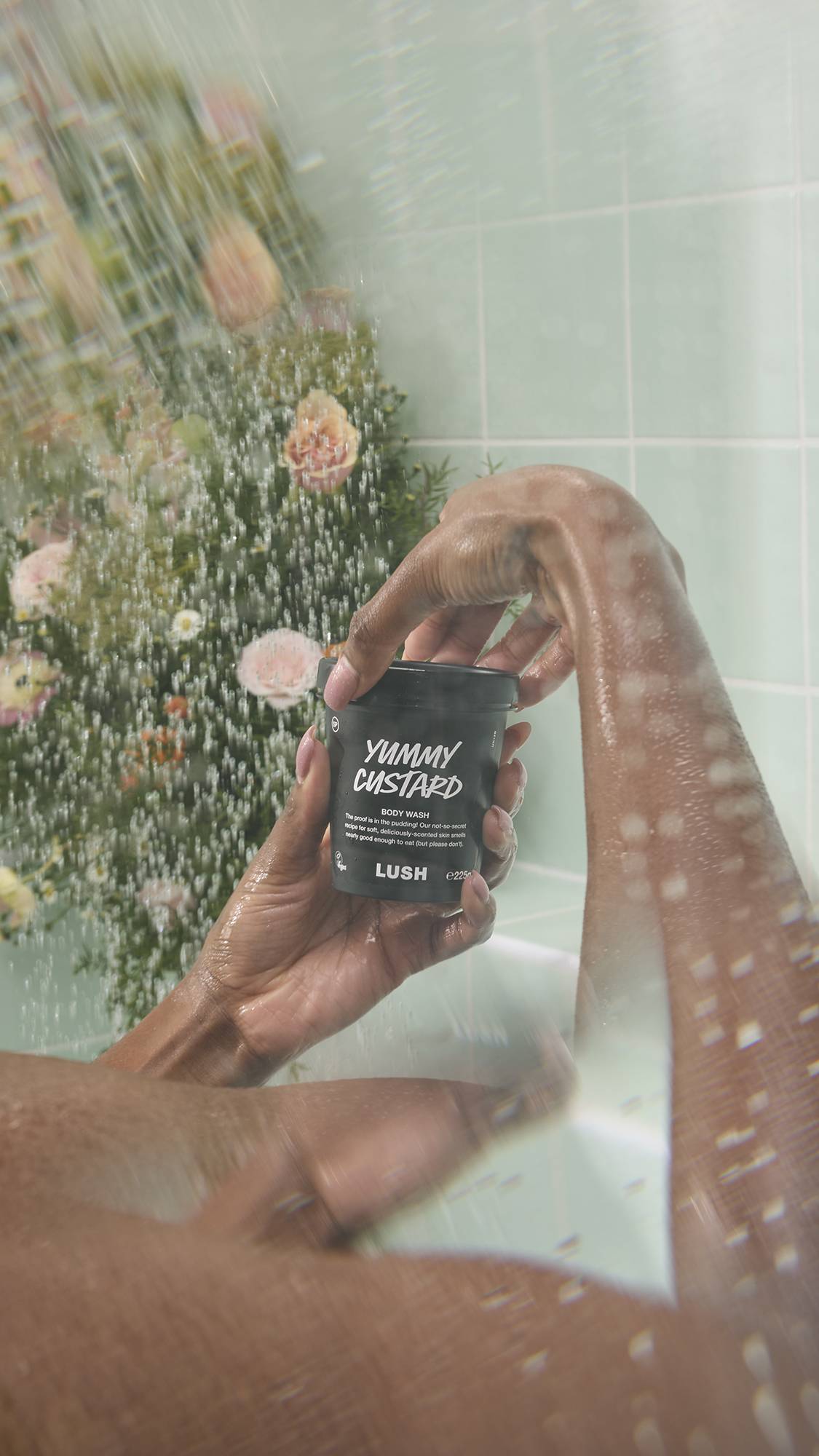 A person holds a black pot of "Yummy Custard" body wash in the shower, surrounded by falling water droplets. The packaging features the text "The proof is in the pudding!" and is set against a backdrop of mint green tiles and blurred flowers.