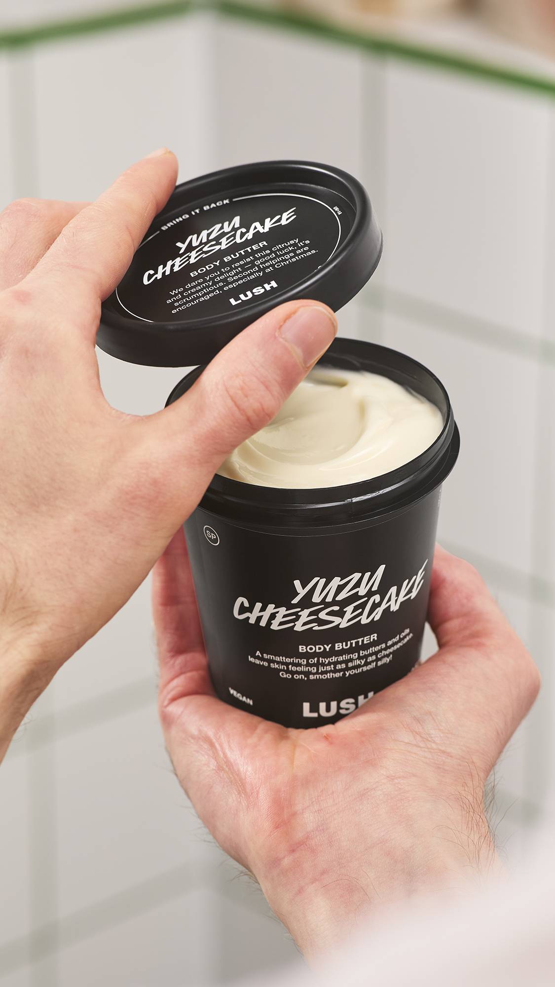Hands hold a black tub of Lush Yuzu Cheesecake body butter, lifting off the black lid to reveal rich cream-coloured body butter inside, set against a white tiled bathroom background.