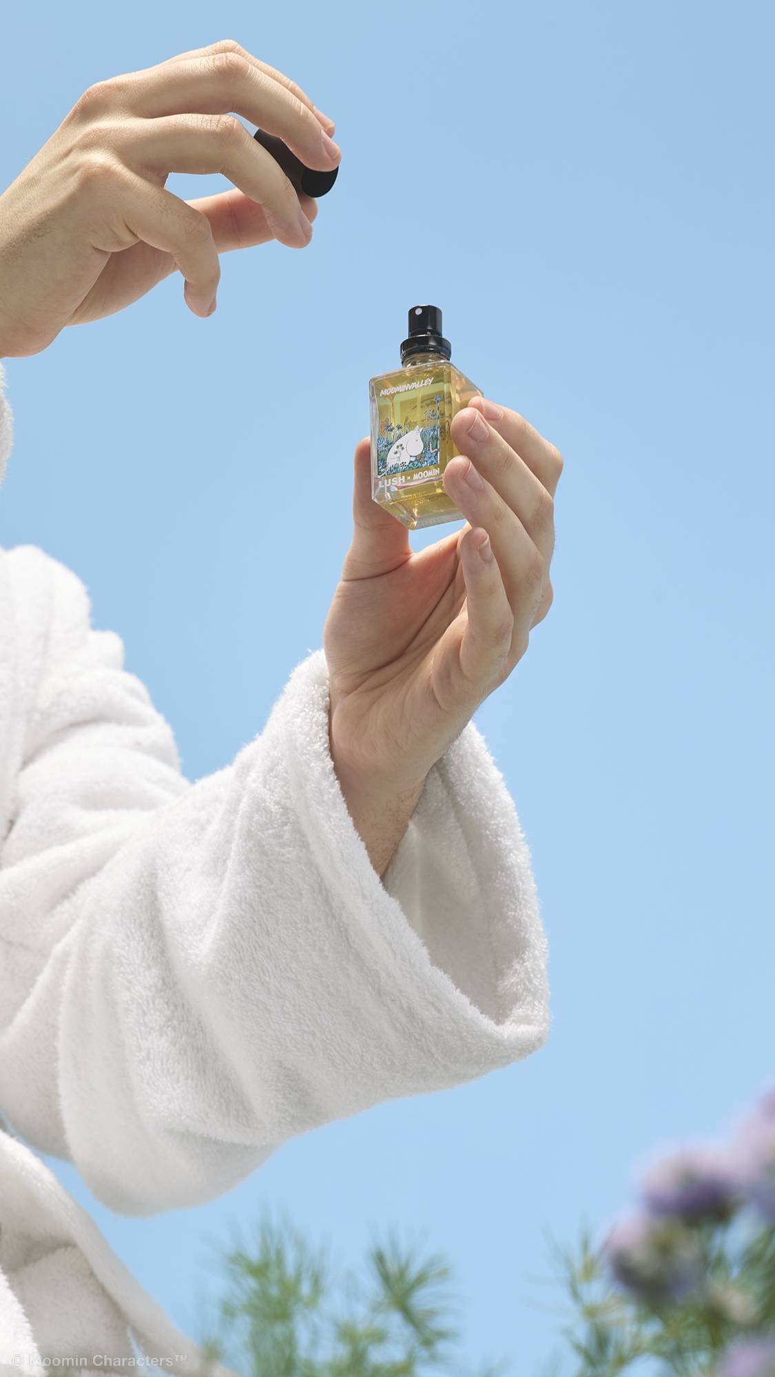 A close-up shot of a person's hands in a white, fluffy dressing gown holding up a small glass bottle of Lush x Moomin perfume against a clear blue sky. One hand gently holds the bottle whilst the other lifts off the black lid.