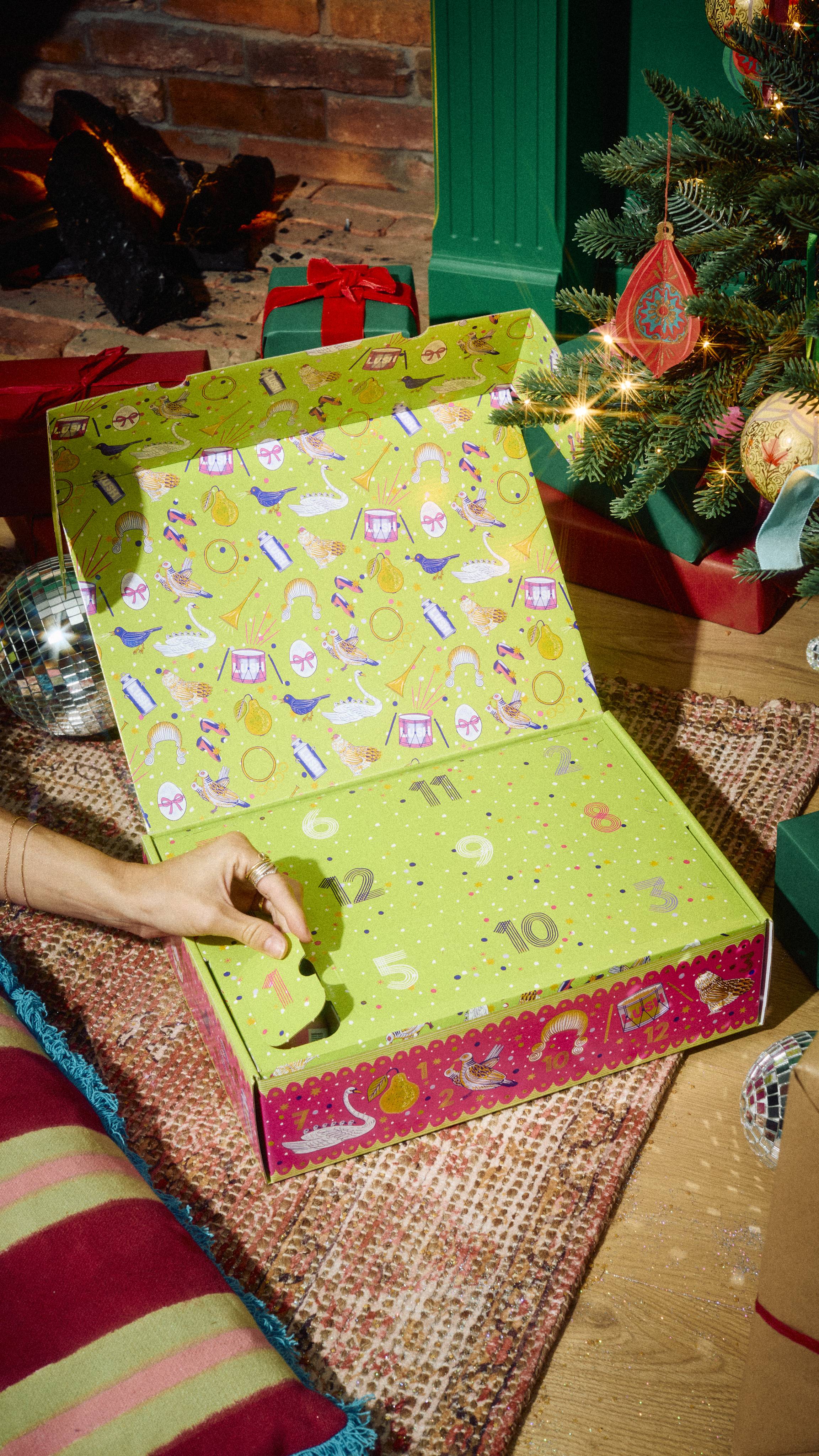 A hand opens door number one on the Lush 12 Days of Christmas countdown box, resting on a rug in a festive room setting by a cozy fireplace and decorated Christmas tree.