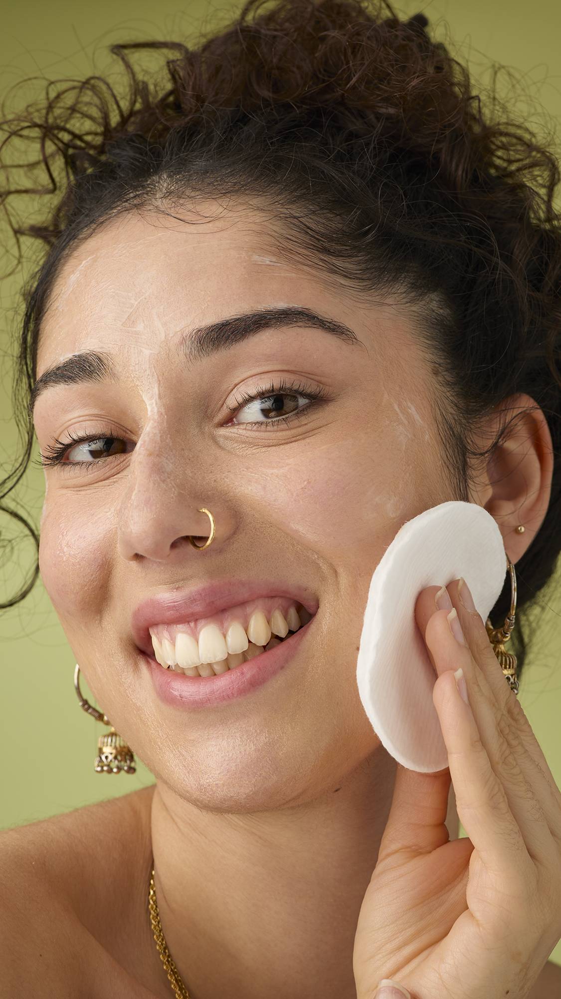 A joyful person smiles broadly while applying the 9 to 5 cleanser with a circular pad to their cheek and nose.