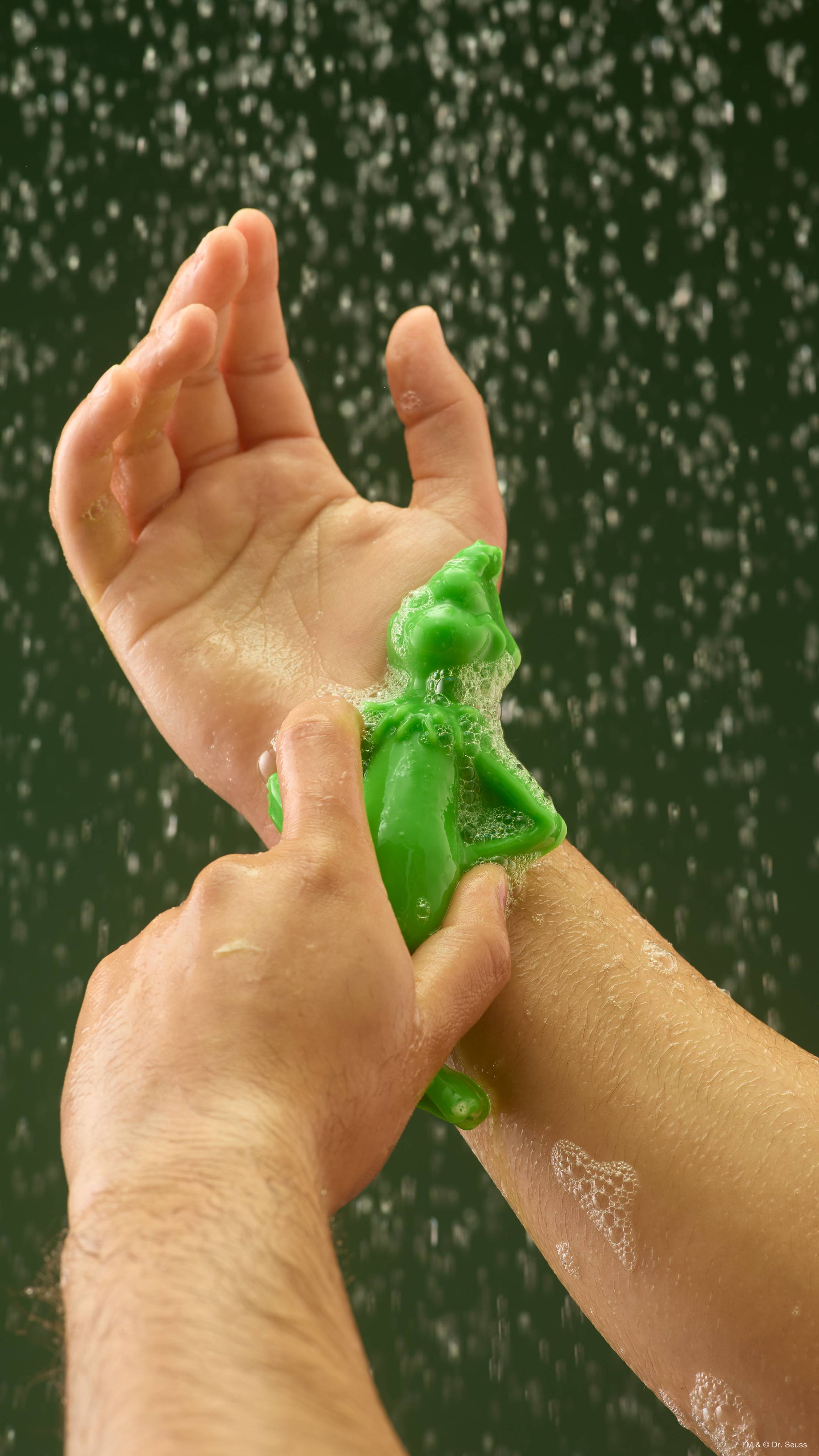 A bright green Lush shower gummy shaped like The Grinch is being rubbed onto a person's wrist, creating white foamy suds under streaming water droplets against a dark green backdrop.  
